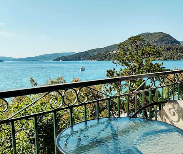 Et glassbord og stoler på en balkong med utsikt mot havet, grøntområder og fjell i bakgrunnen. (AI-generert tekst)