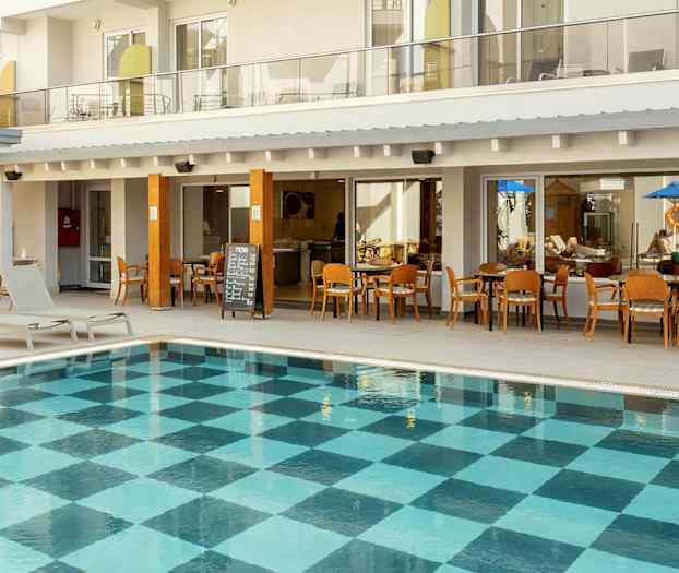 Utendørsbasseng med rutet bunnmønster foran hotellbygning med balkonger og restaurant. (AI-generert tekst)