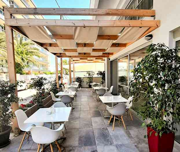 Uteservering ved en bygning med bord, stoler og tak av tre med duker. (AI-generert tekst)