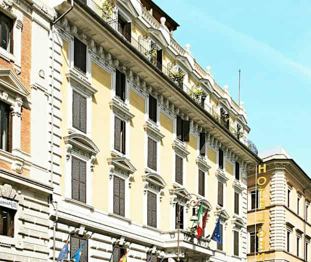 Fasaden til et hotell med flere etasjer, dekorert med flagg og omgitt av biler og motorsykler. (AI-generert tekst)