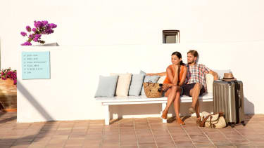 Par på ferie sitter på en benk med koffert ved siden av seg