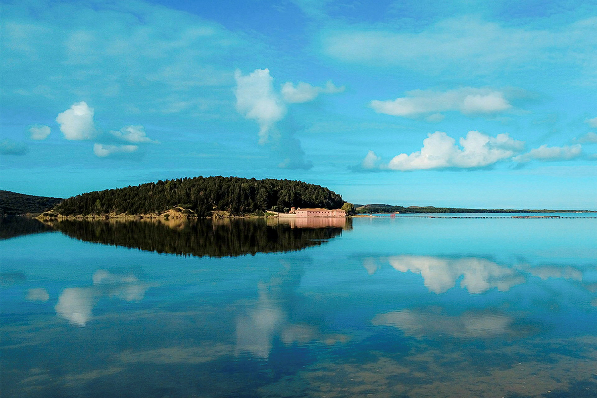 En øy med skog og bygninger omgitt av rolig vann, under en klar himmel med skyer. (AI-generert tekst)