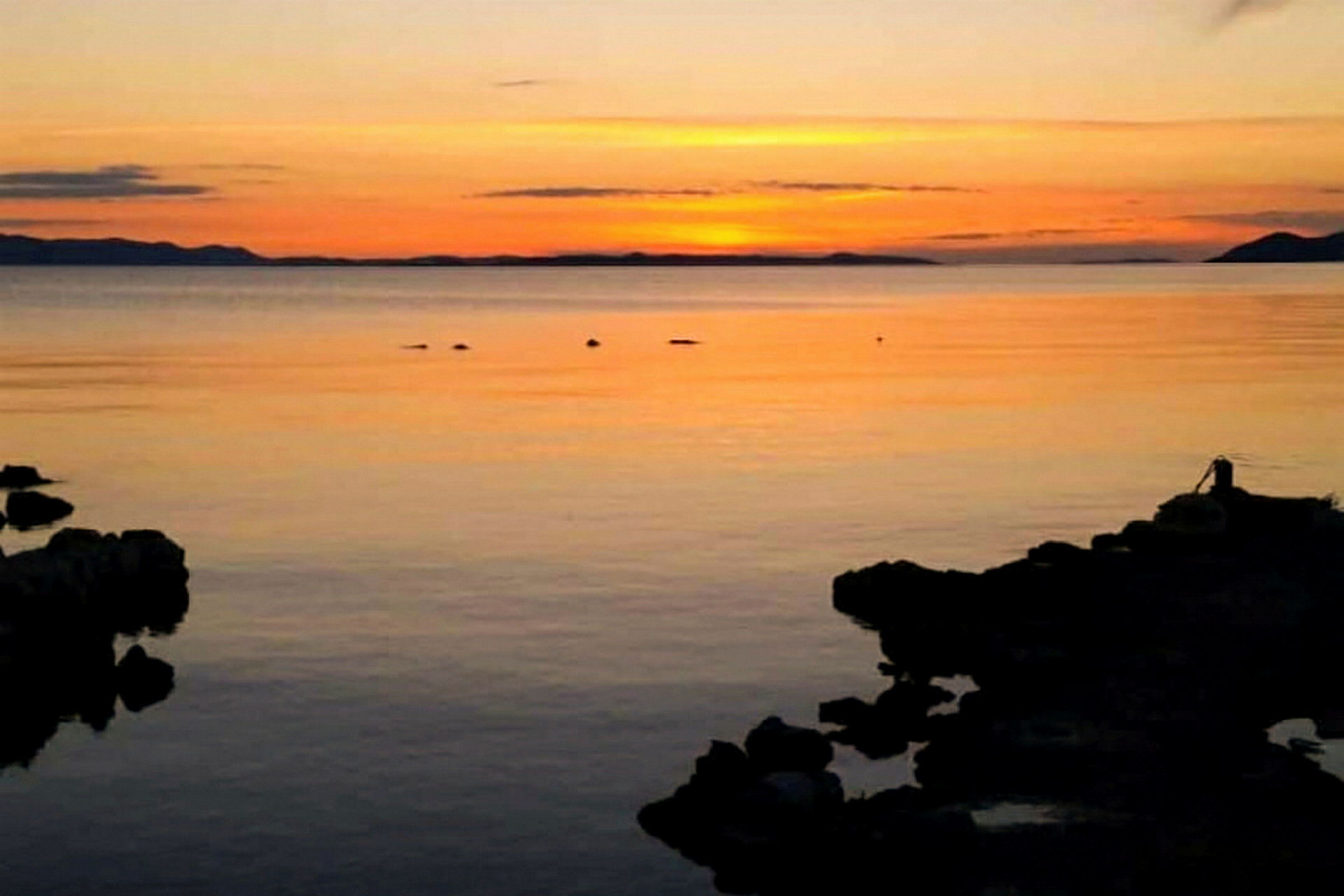 Solnedgang over et rolig hav med silhuetter av klipper i forgrunnen. (AI-generert tekst)