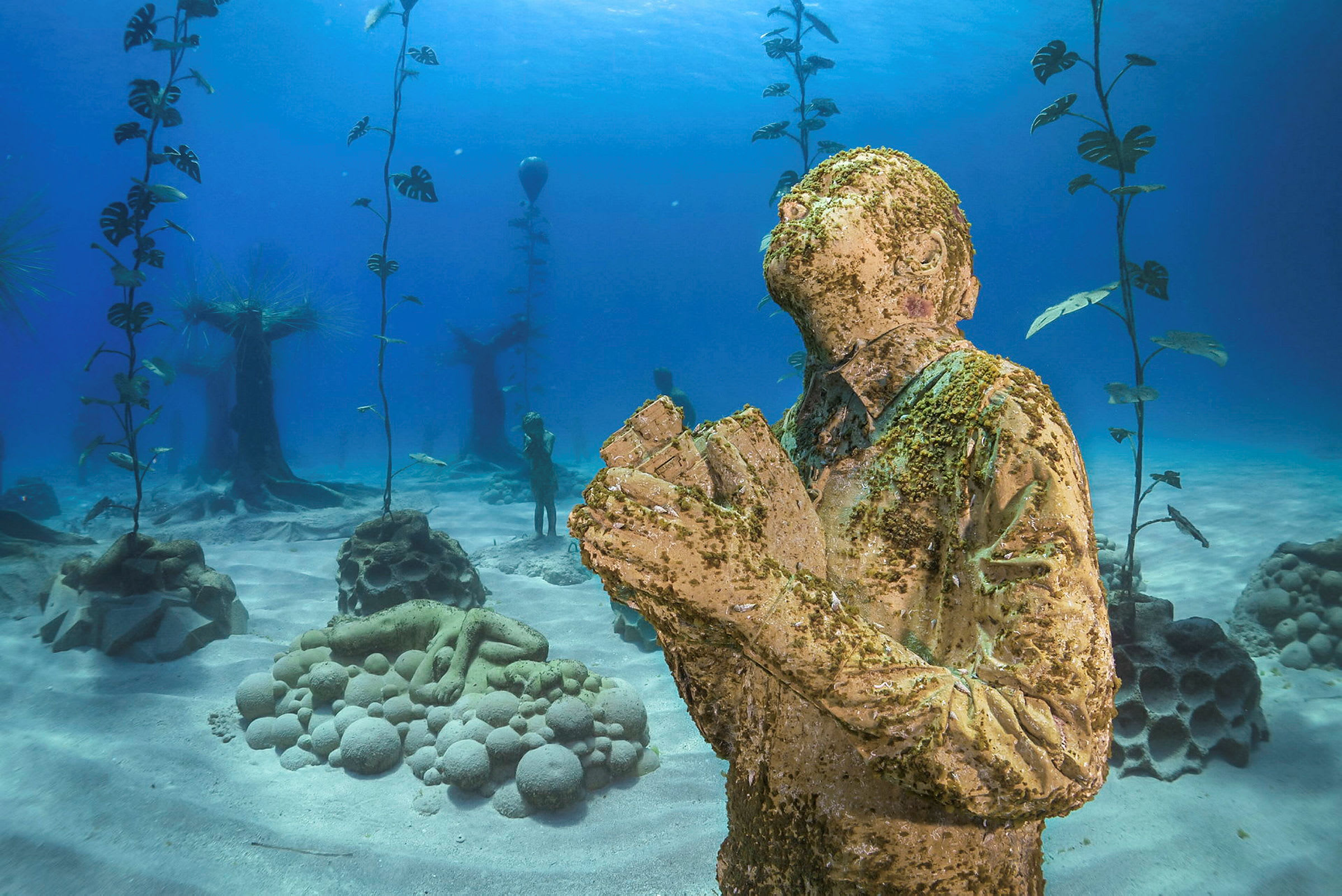 Undervannsbilde som viser en statue av en person i en bønnestilling, omgitt av undervannsplanter og andre marine skulpturer. (AI-generert tekst)