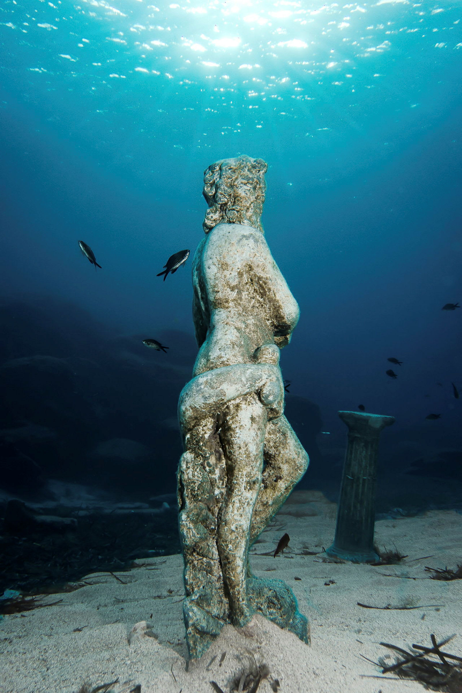 Et undervannsbilde av en gammel statue omgitt av sand og småfisk. (AI-generert tekst)