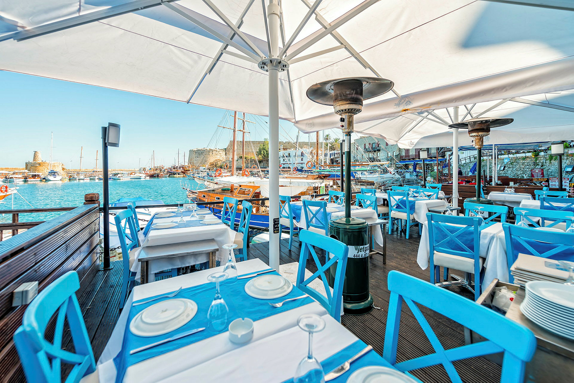 En restaurant med blå stoler og hvite bord ved en havn. Seilbåter er synlige i bakgrunnen. (AI-generert tekst)