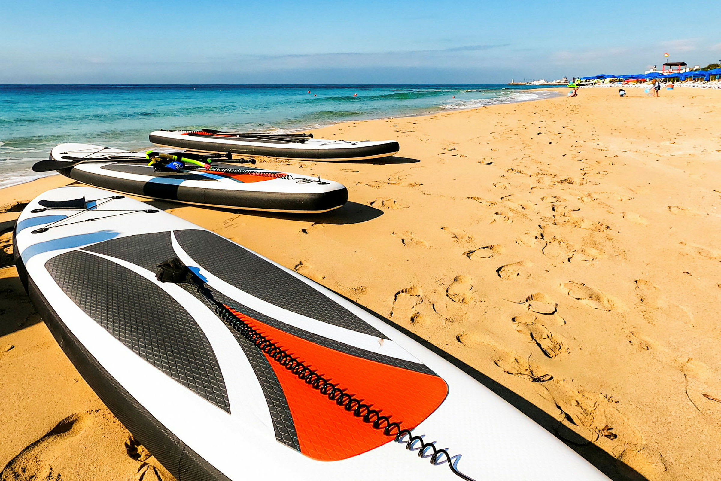 Bilde av en sandstrand med flere stand up paddleboards plassert på sanden nær vannet. (AI-generert tekst)
