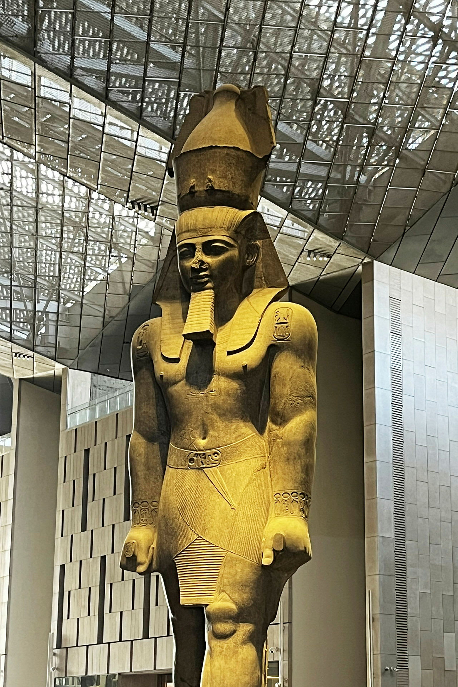 En stor antikk statue av en stående mannsfigur med detaljert antrekk og hodeplagg, plassert i en moderne bygning. (AI-generert tekst)