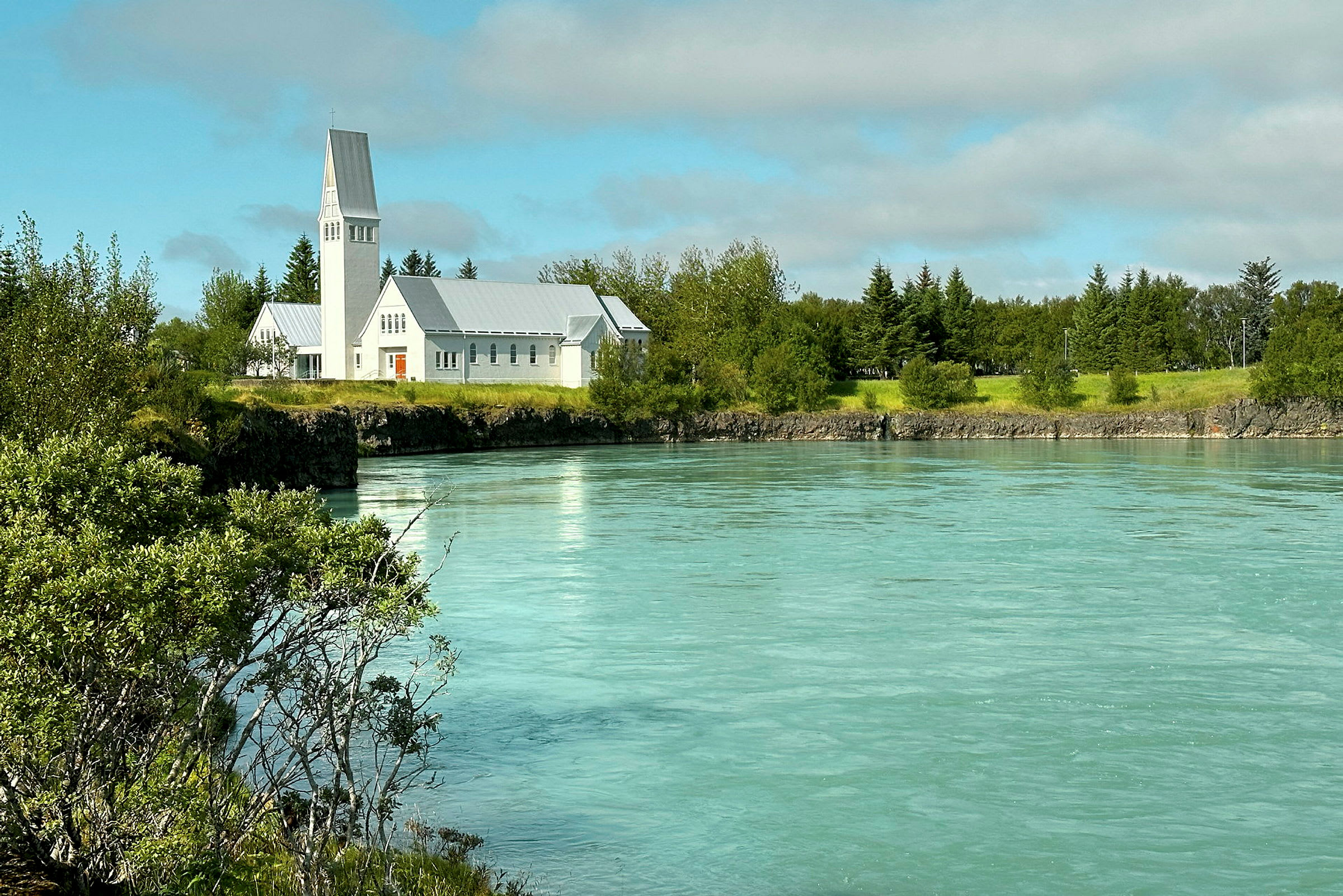 En hvit kirke ligger ved en turkisfarget elv omgitt av grøntområder og trær. (AI-generert tekst)