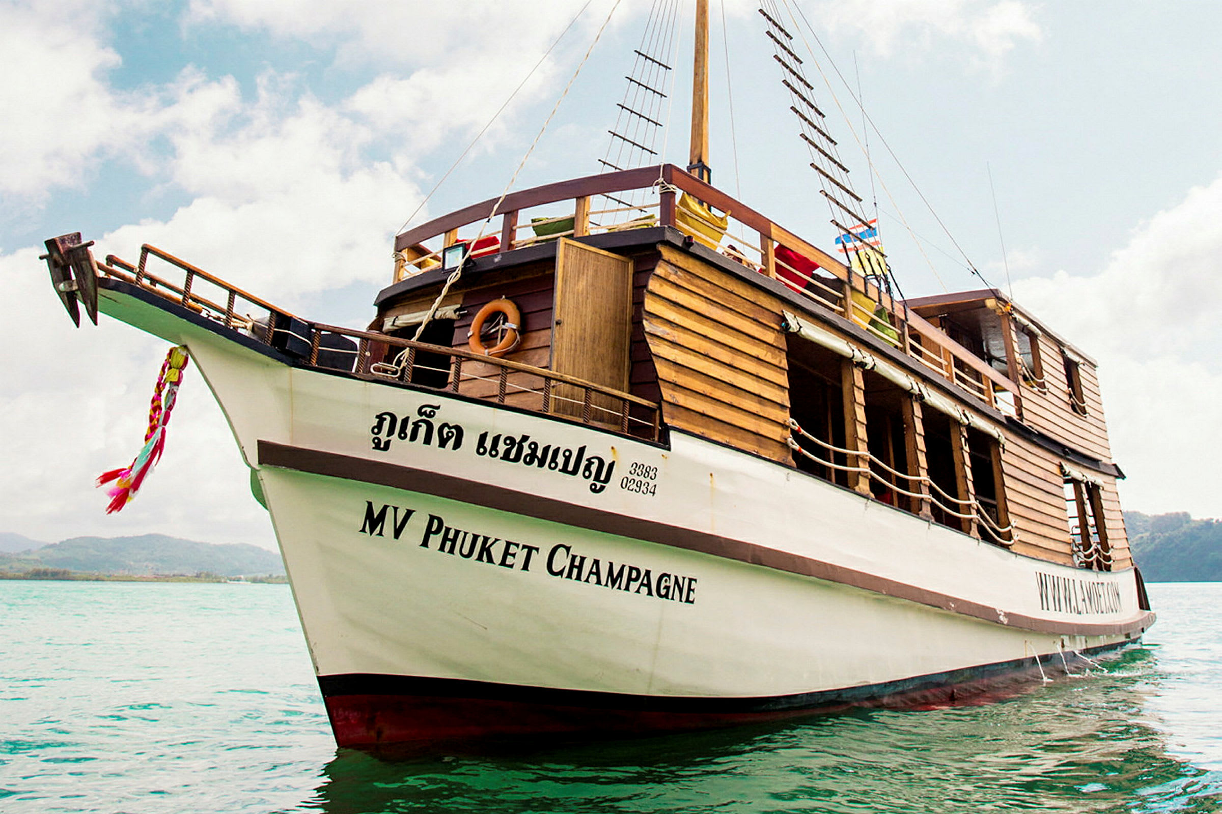 Et trefartøy med teksten 'MV Phuket Champagne' på skroget ligger forankret i klart vann. (AI-generert tekst)
