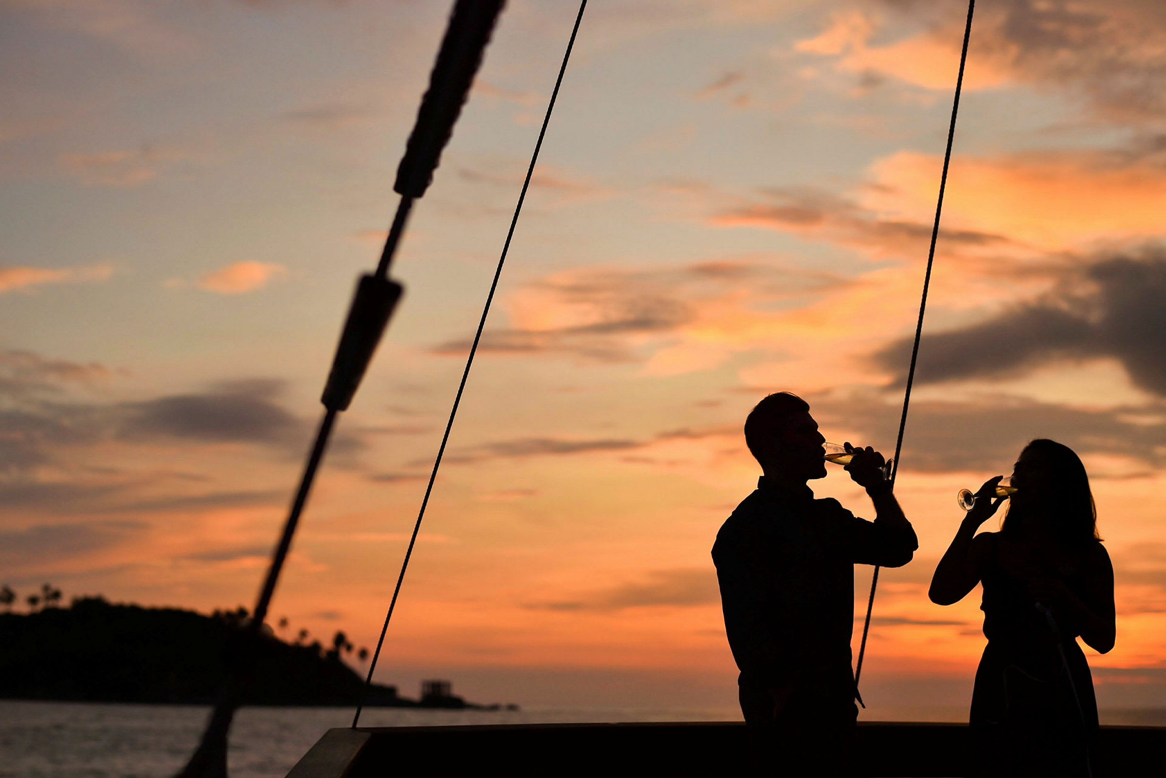 Silhuetter av to personer som holder glass ved solnedgang med havet og en liten øy i bakgrunnen. (AI-generert tekst)
