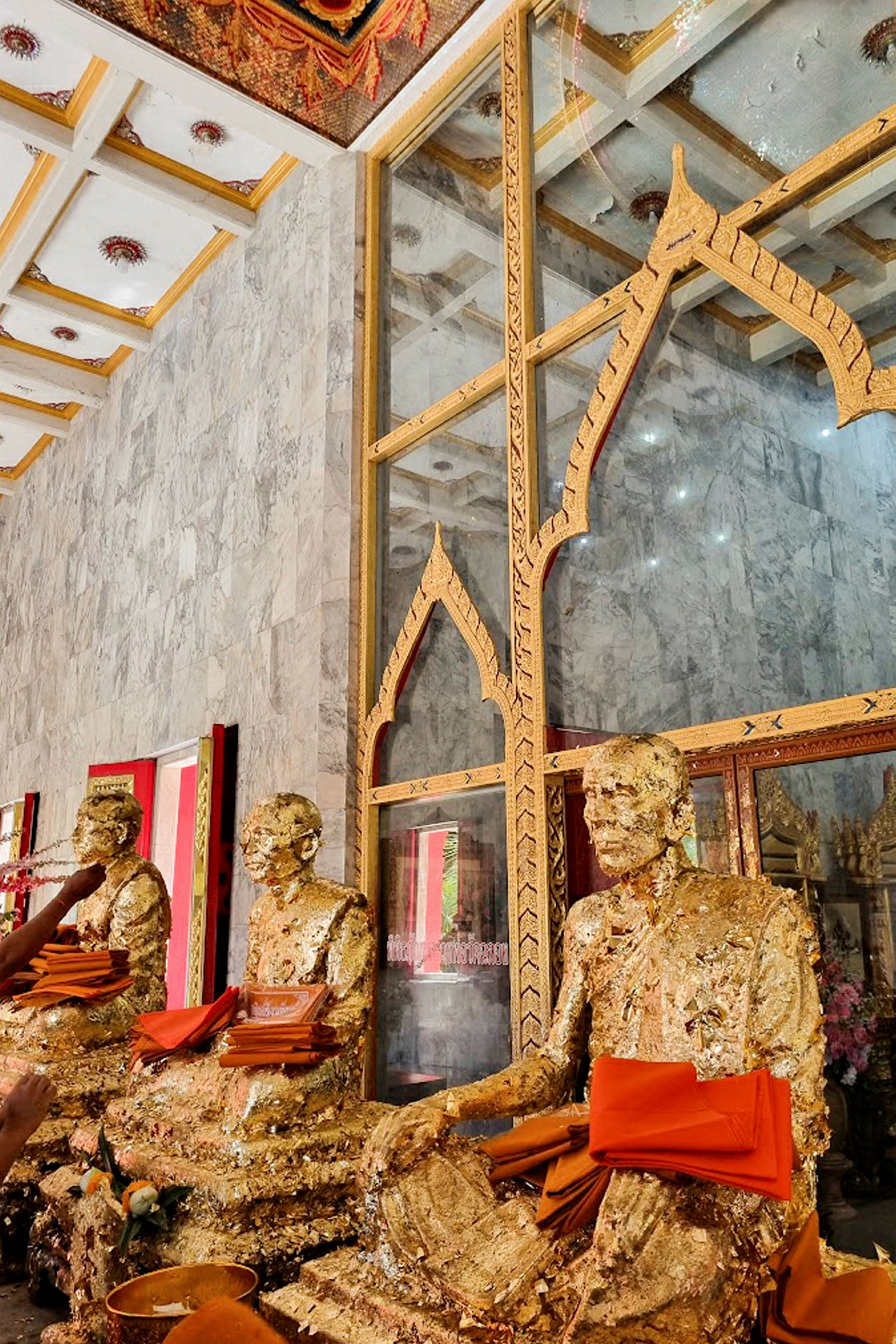 Bilde av gullfargede buddhistiske statuer dekorert med oransje tekstiler foran en glassvegg med utsmykninger. (AI-generert tekst)