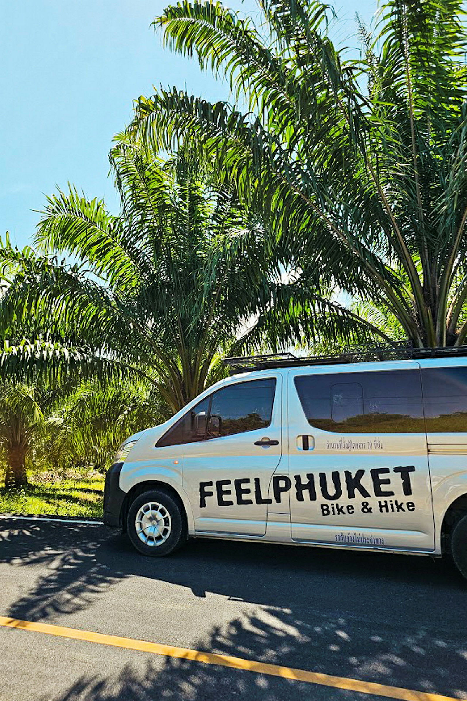 En minibuss med teksten 'Feel Phuket Bike & Hike' parkert langs veien nær palmer. (AI-generert tekst)