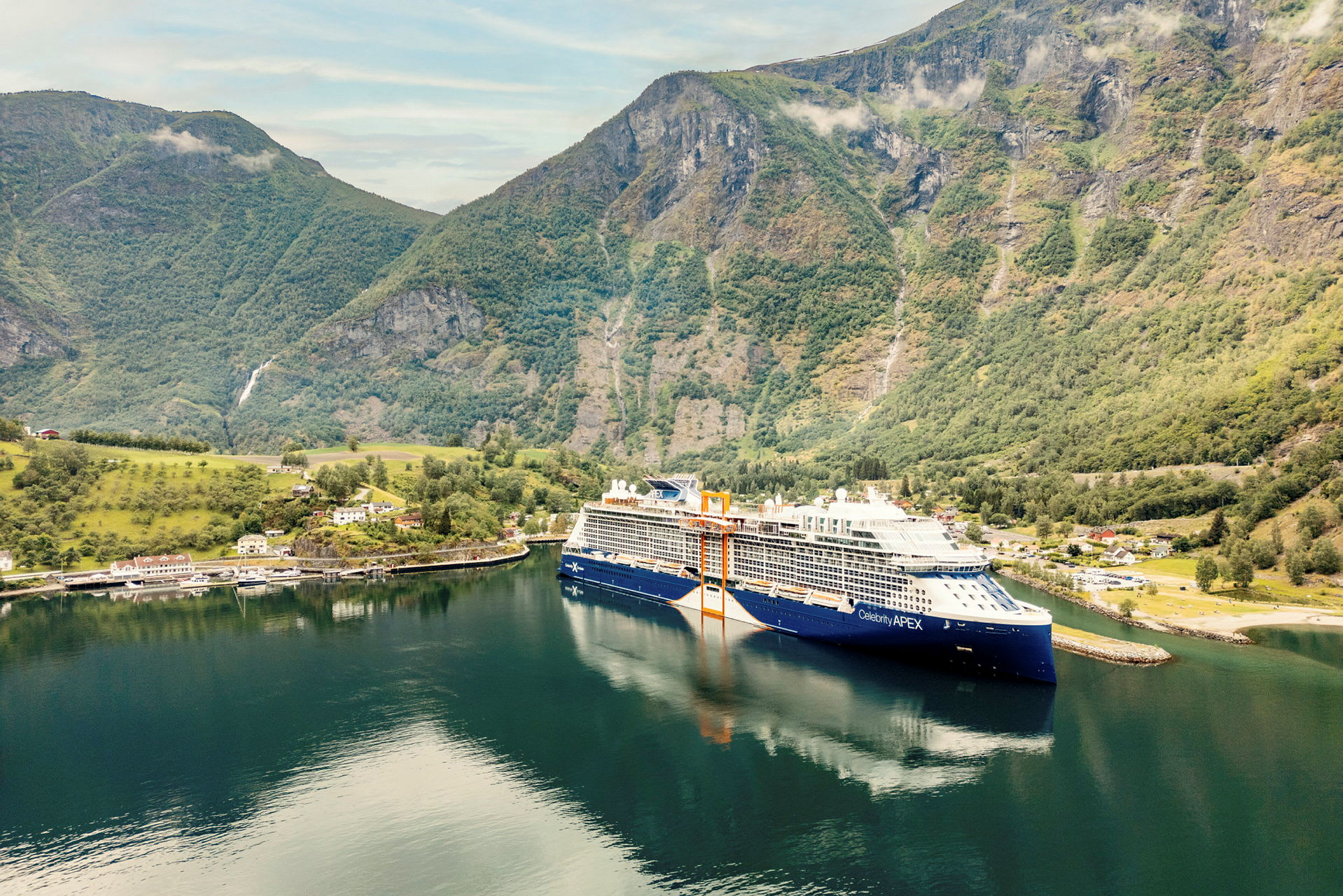 Et stort cruiseskip ligger til kai ved en havn omgitt av høye fjell og grøntområder. (AI-generert tekst)