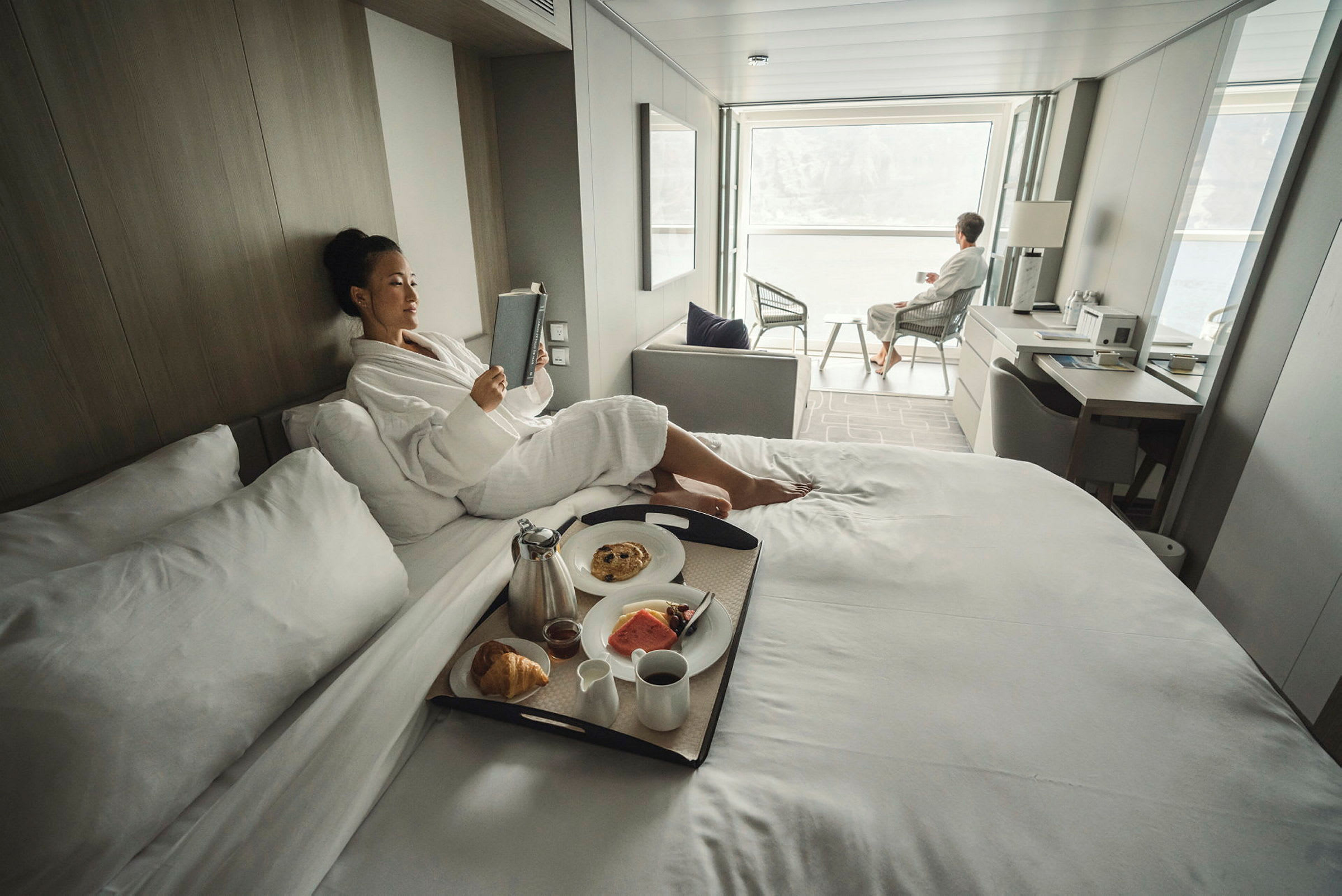 Personer slapper av på et lyst og moderne hotellrom, med frokost servert på sengen og sitteplass ved en stor vindusutsikt. (AI-generert tekst)