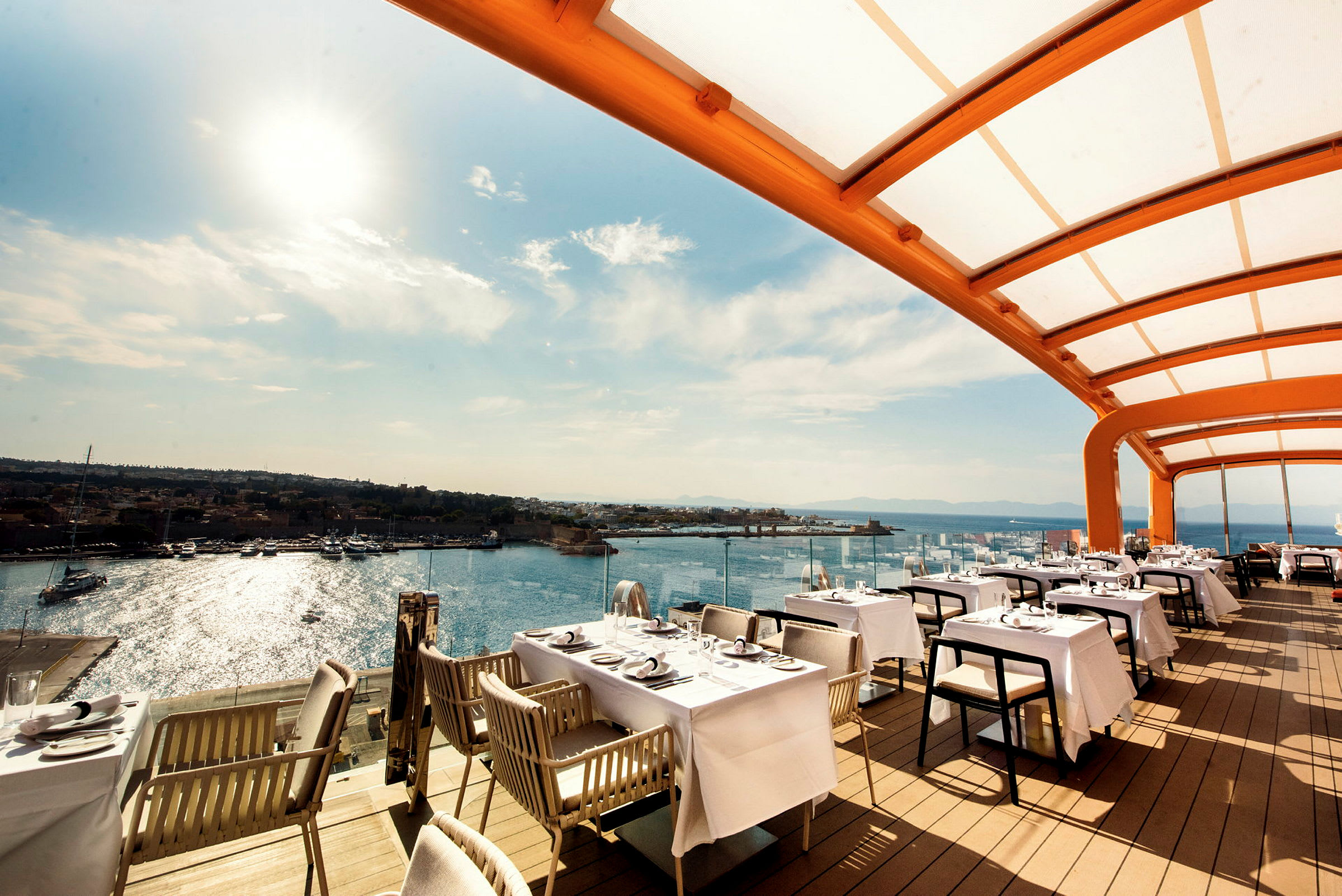 Bilde av en restaurant med utendørs servering under et overbygg, med havet og noen båter i bakgrunnen. (AI-generert tekst)