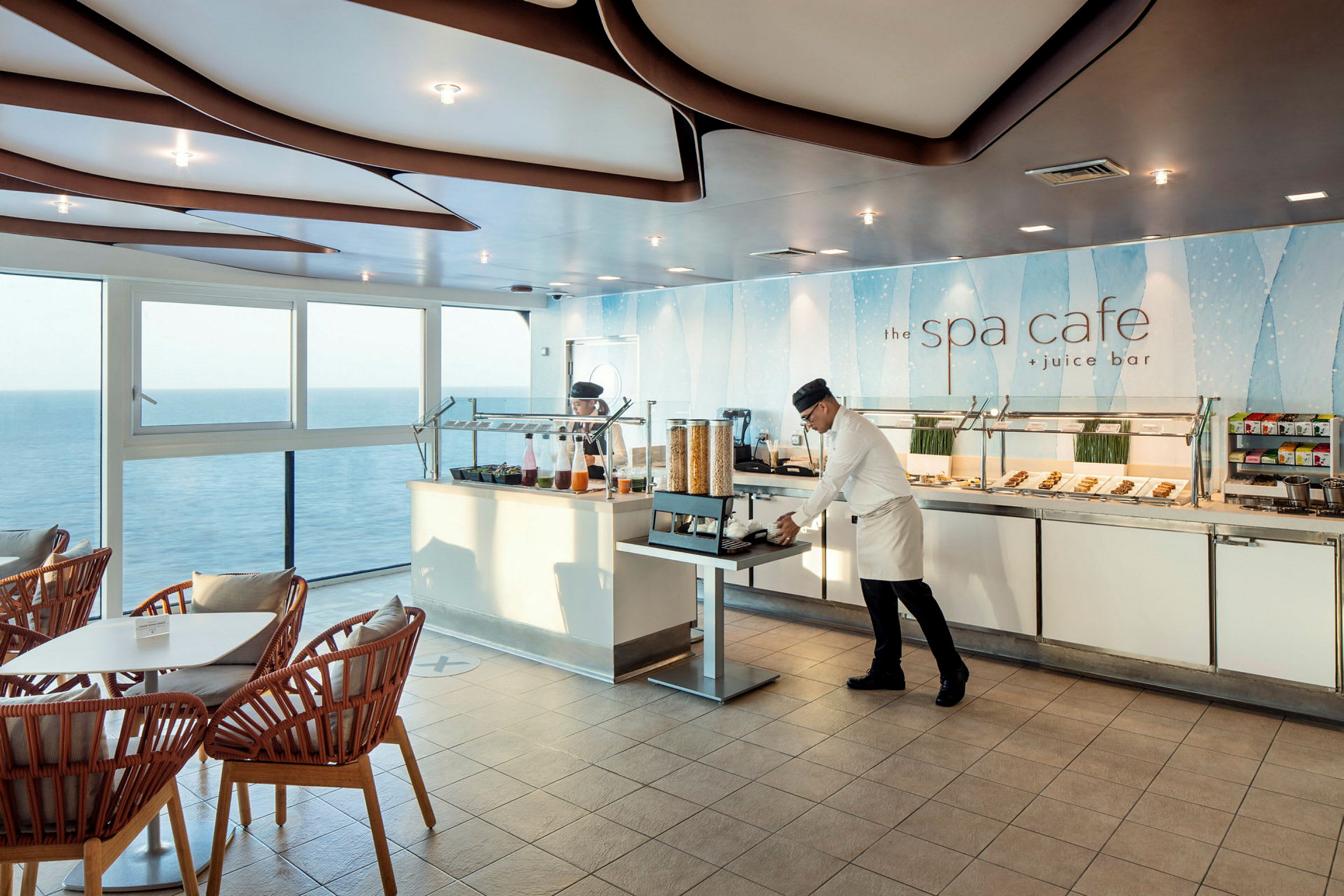 En kafé med store vinduer som gir utsikt over havet, med sitteplasser og en buffet fylt med mat og drikke. (AI-generert tekst)