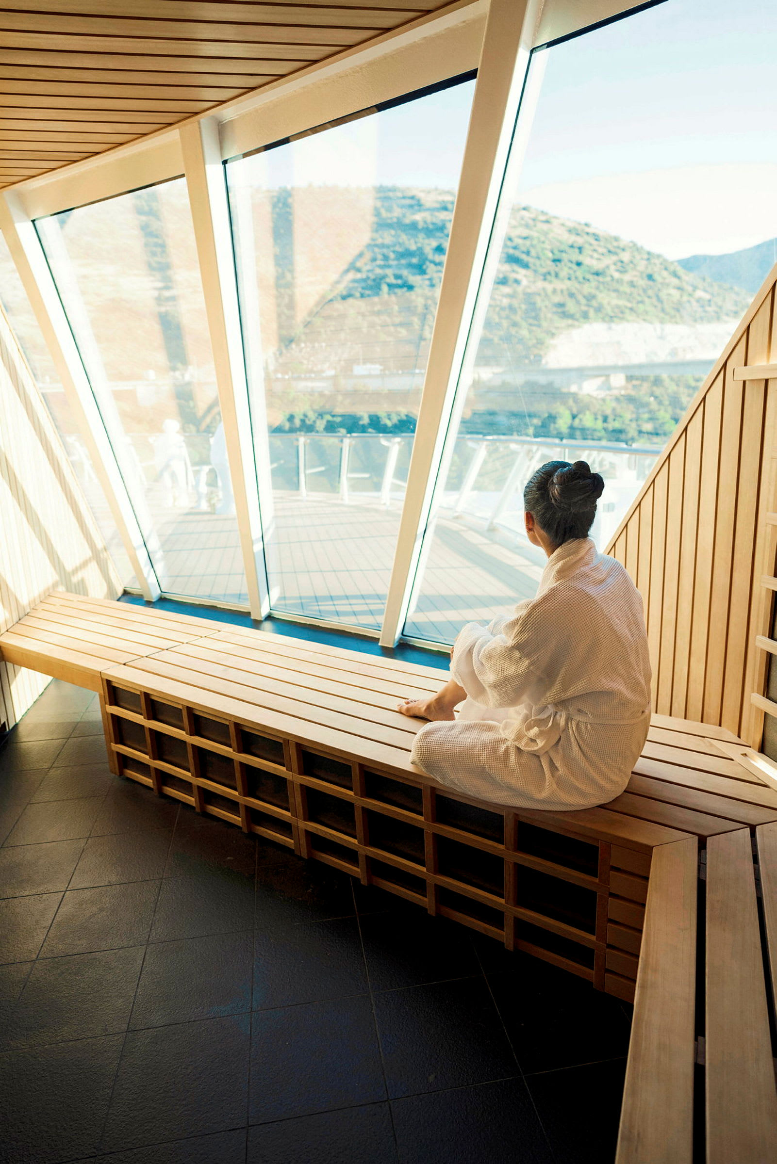 En person i en hvit badekåpe sitter på en benk innendørs med en stor glassvegg som gir utsikt over et fjellandskap utenfor. (AI-generert tekst)