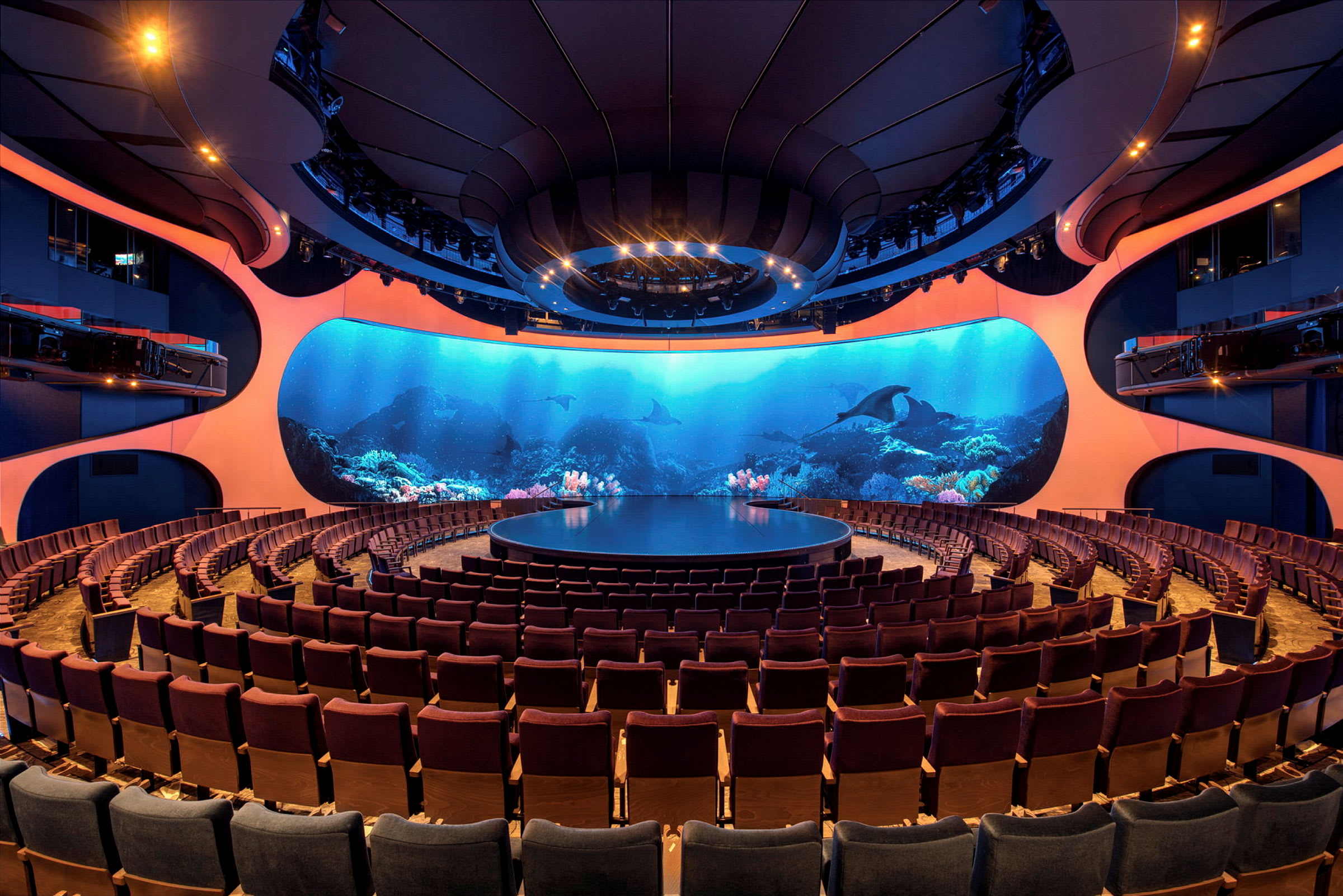 Et auditorium med en rund scene og rader med publikumsstoler. Bakgrunnen består av en stor skjerm som viser et undervannsmotiv. (AI-generert tekst)