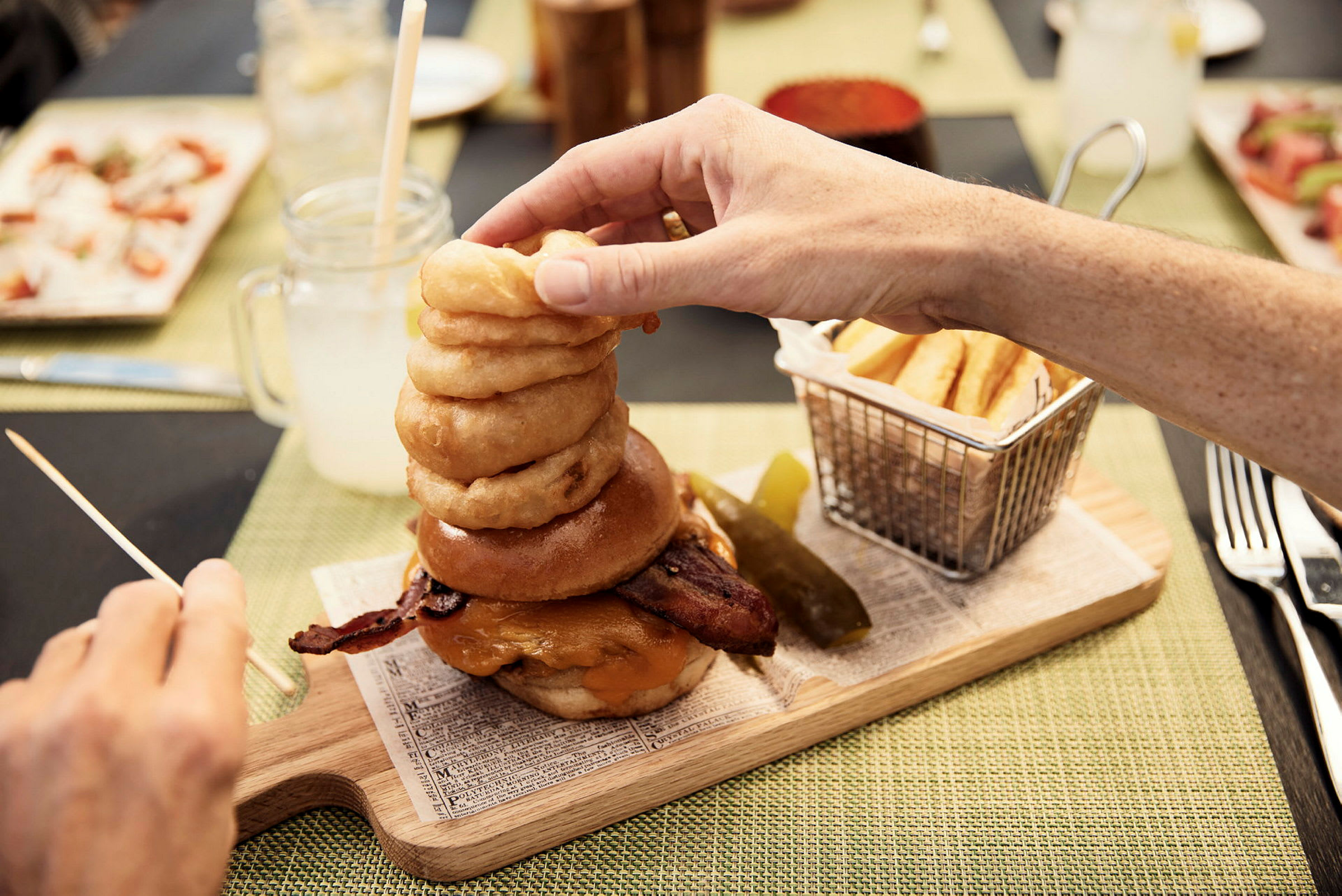 En hamburger med bacon, ost og løkringer, servert med pommes frites og en agurkbit. (AI-generert tekst)