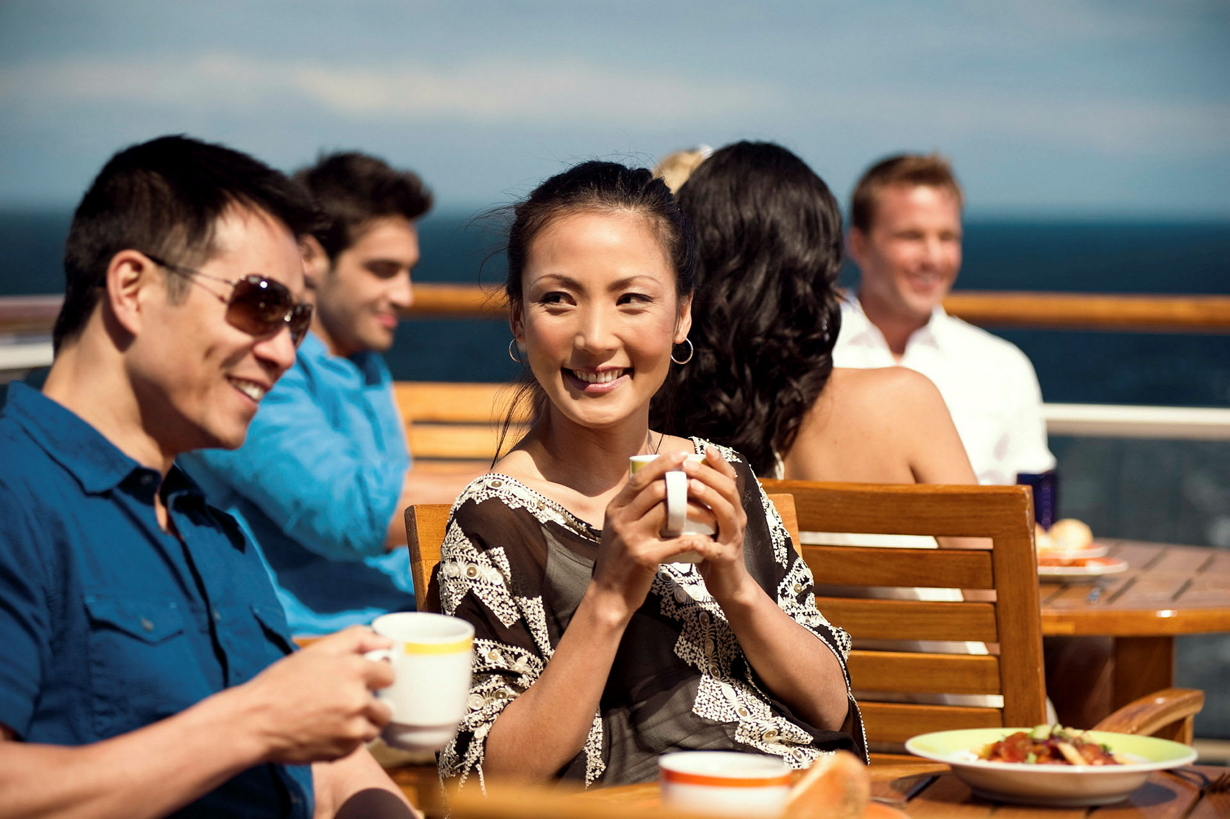Personer sitter ved et bord og drikker varme drikker på en terrasse med havutsikt. (AI-generert tekst)