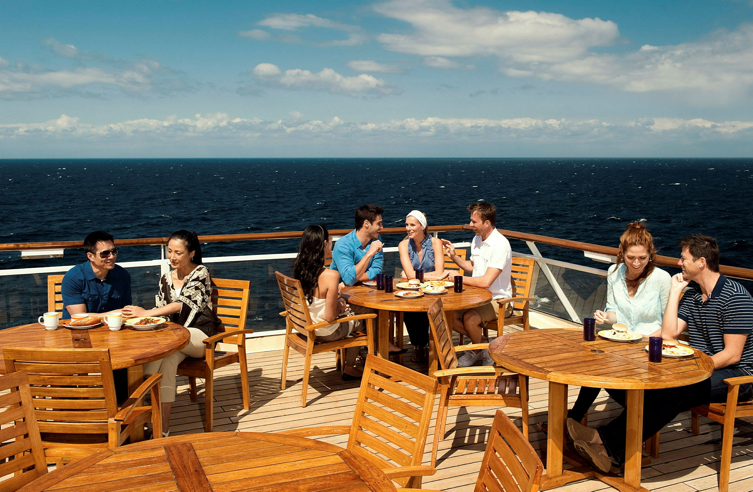Personer sitter ved tremøbler på en utendørs terrasse med utsikt over havet. (AI-generert tekst)