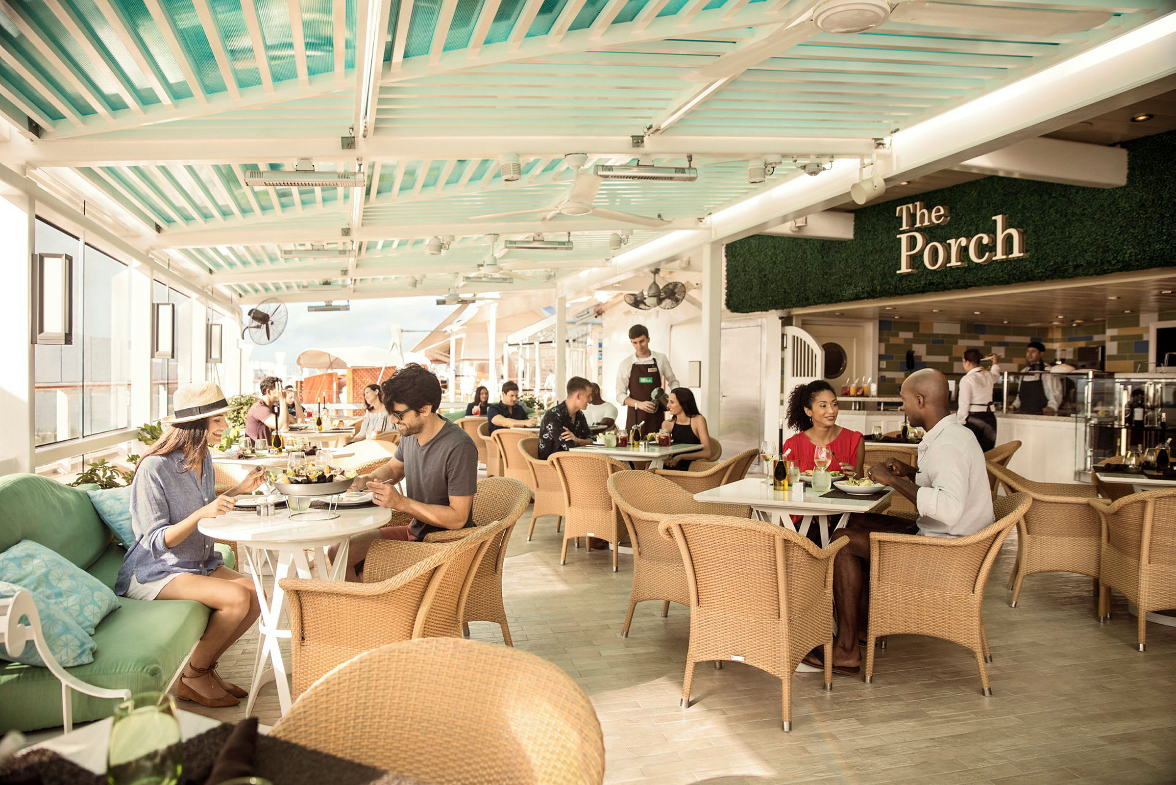 En restaurant med gjester som sitter ved bord og spiser og prater. Innredningen har rottingstoler og et soltak. (AI-generert tekst)