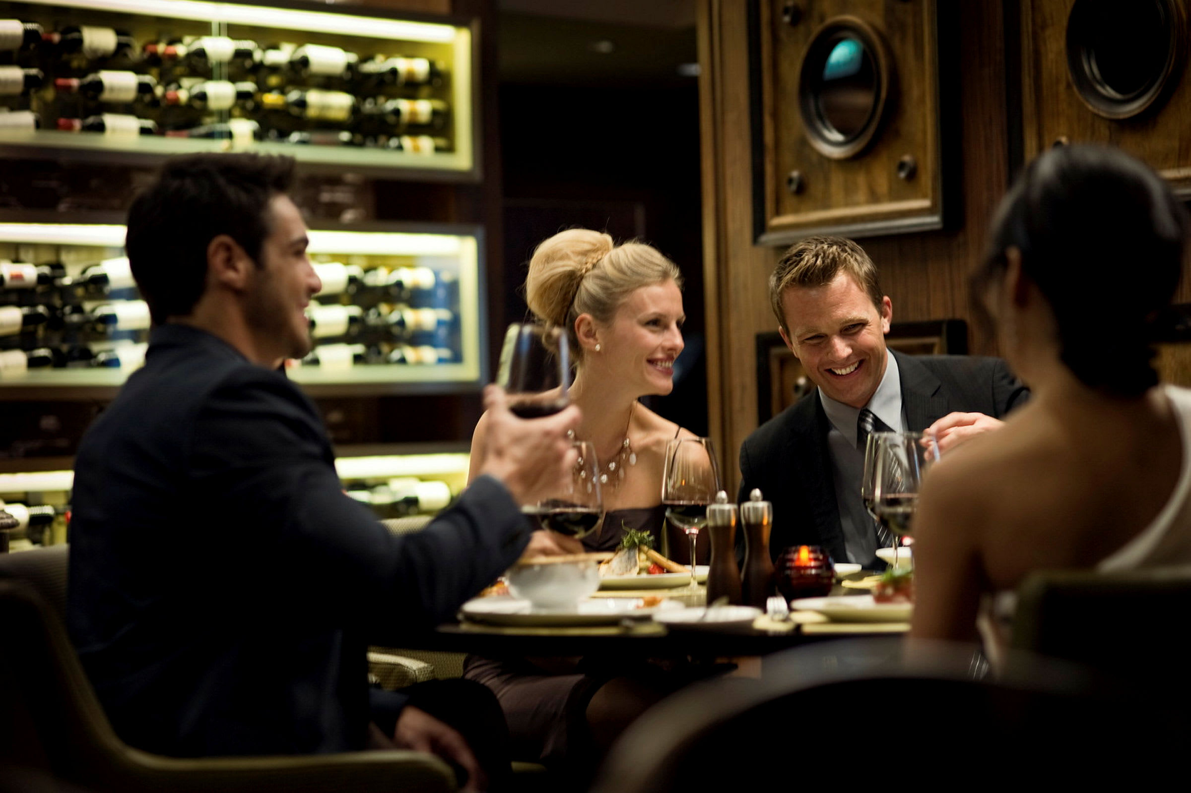 En gruppe mennesker nyter mat og vin ved et bord i en elegant restaurant med en vegg fylt med vinforsyninger i bakgrunnen. (AI-generert tekst)