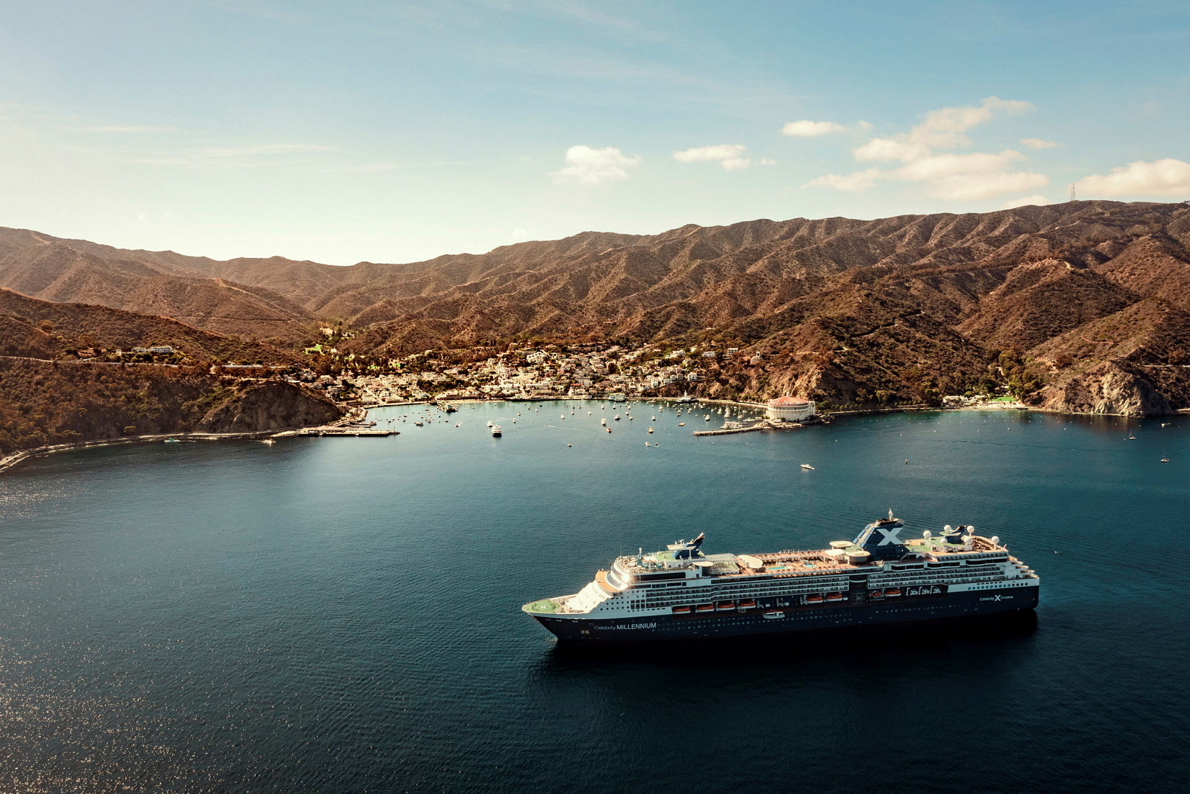 Et stort cruiseskip ligger i en bukt nær en by omgitt av fjell. (AI-generert tekst)