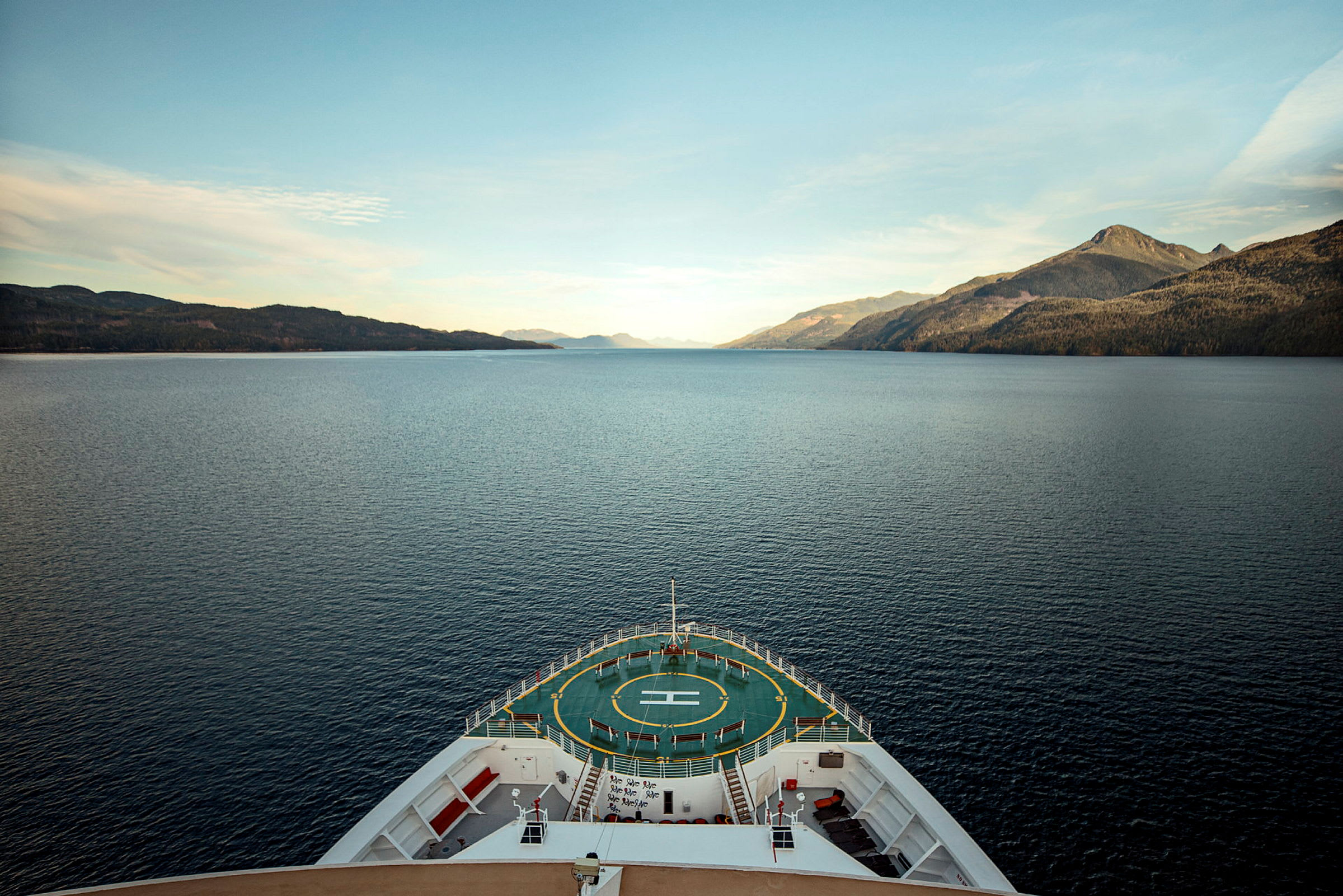 Utsikt fra cruiseskipets baug over en rolig fjord med omkringliggende fjell. (AI-generert tekst)