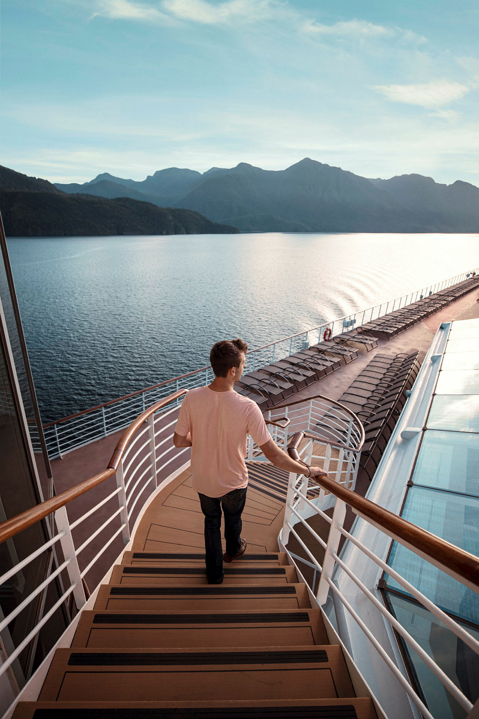 En person går ned en trapp på et cruiseskip, med utsikt over sjøen og fjellene i bakgrunnen. (AI-generert tekst)