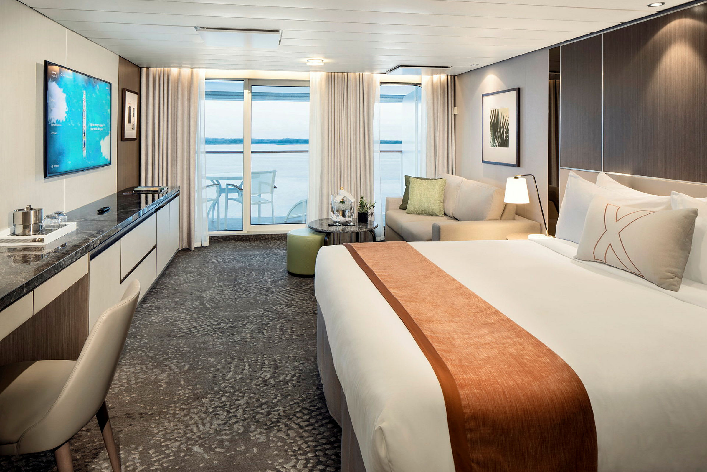 Et moderne hotellrom med en stor seng, sofa og et bord, samt utsikt gjennom glassdører mot en balkong og havet. (AI-generert tekst)