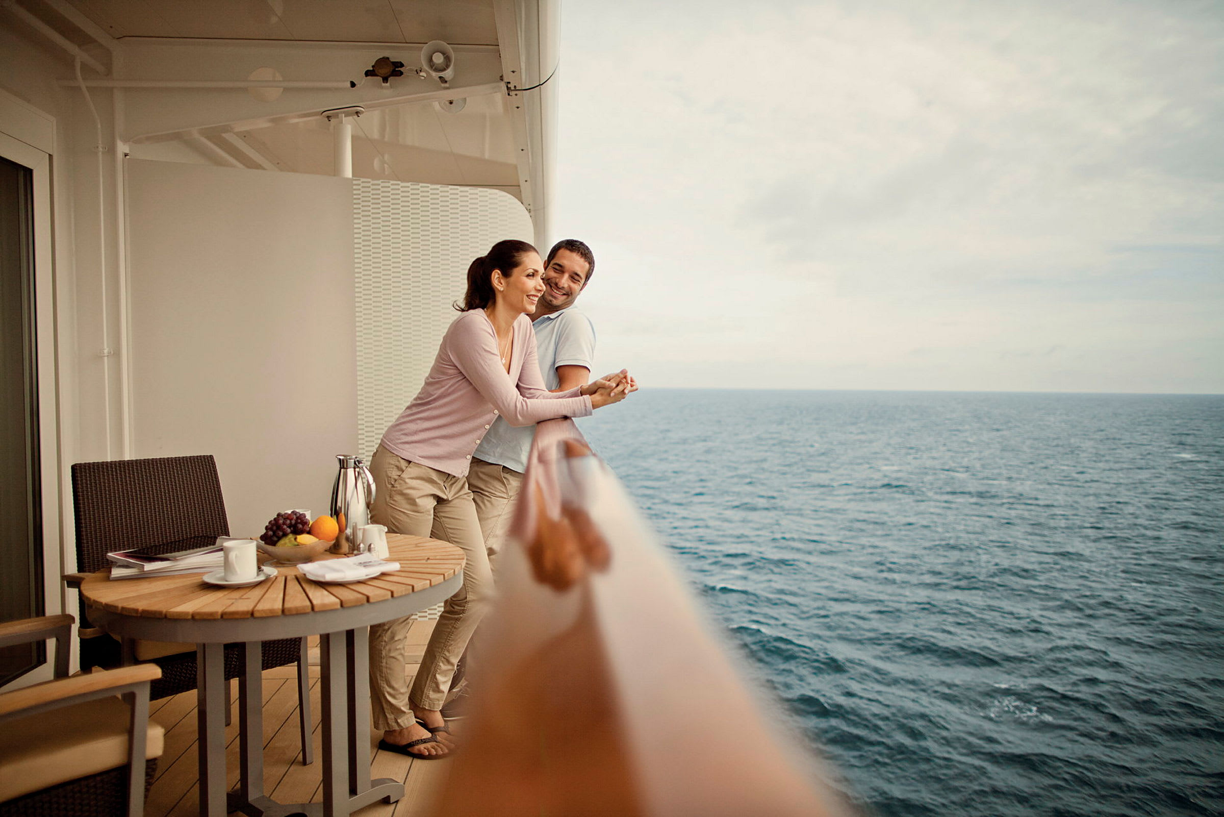 Personer står på en balkong med utsikt over havet. Et trebord er plassert med frukt og drikke. (AI-generert tekst)