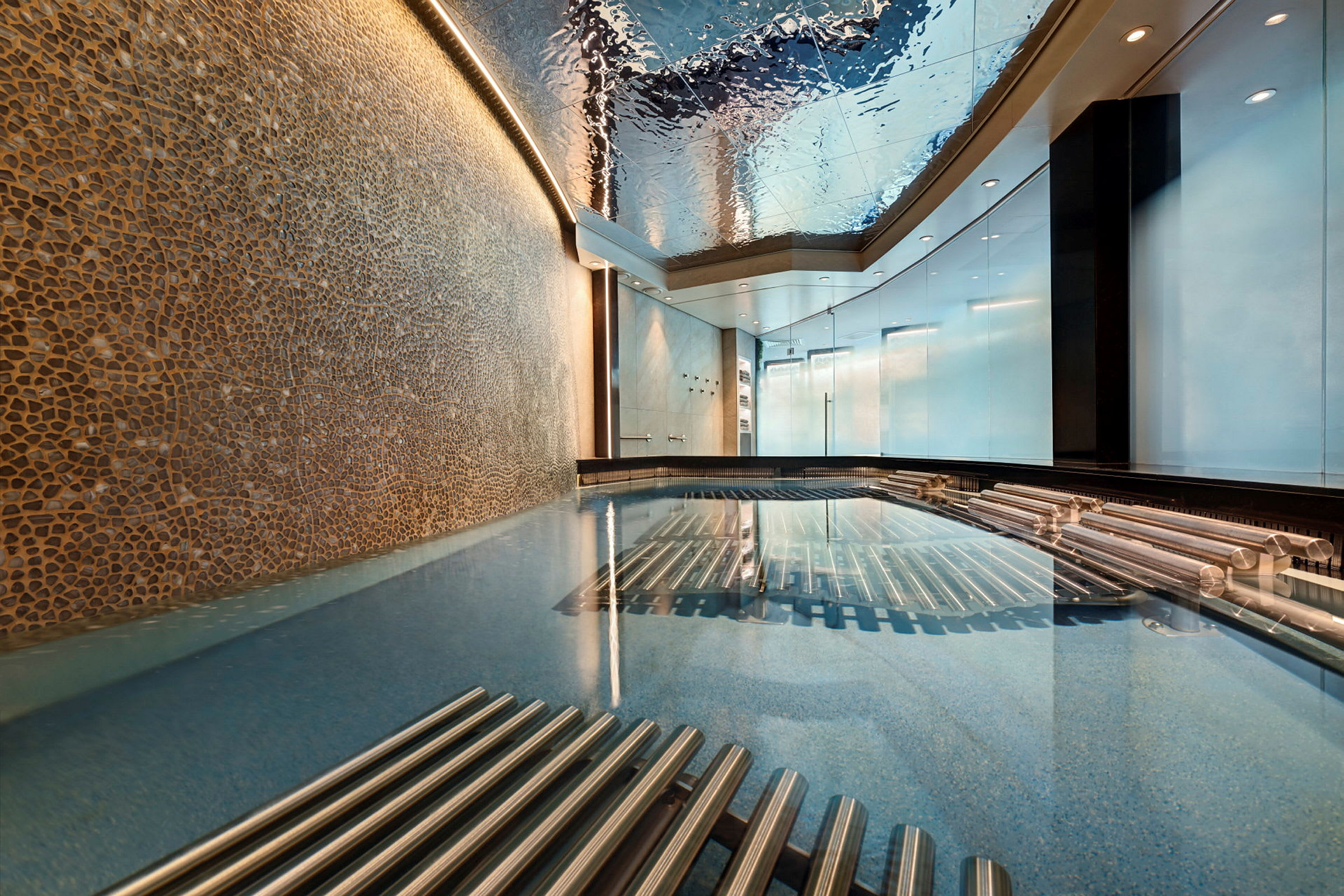 Et innendørs basseng med et tak som reflekterer lys og omgivelser, flankert av metalliske detaljer og vegger med dekorative mosaikker. (AI-generert tekst)