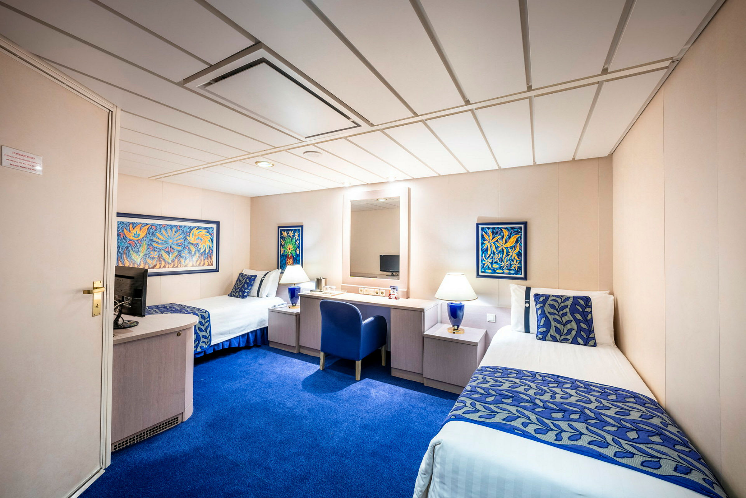 Hotellrom med to enkeltsenger, blå innredning og bord mellom sengene. Veggdekor i farger er tilstede. (AI-generert tekst)