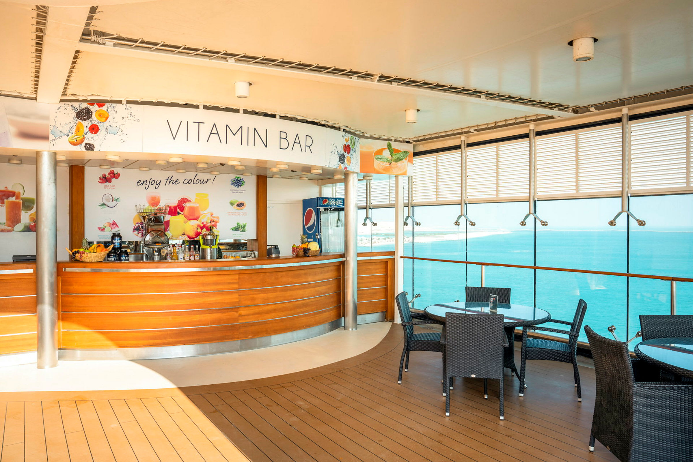 Bilde av en vitaminbar med trepaneler, fargerike frukter som dekorasjon og en glassvegg vendt mot vannet. Noen bord med glassoverflater og mørke rottingstoler vises i forgrunnen. (AI-generert tekst)