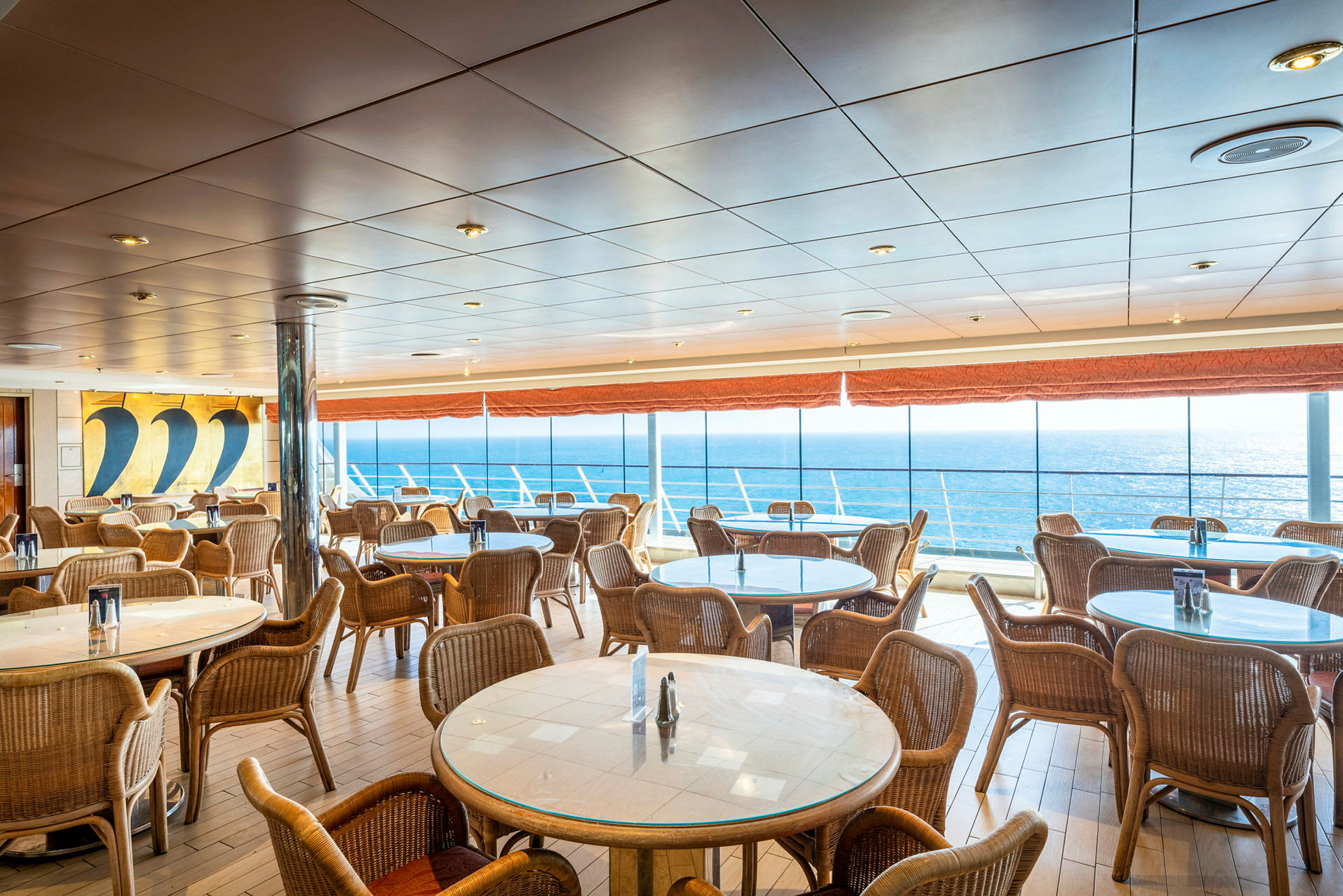 Spiseområde med runde bord og stoler, samt store vinduer med utsikt over havet. (AI-generert tekst)