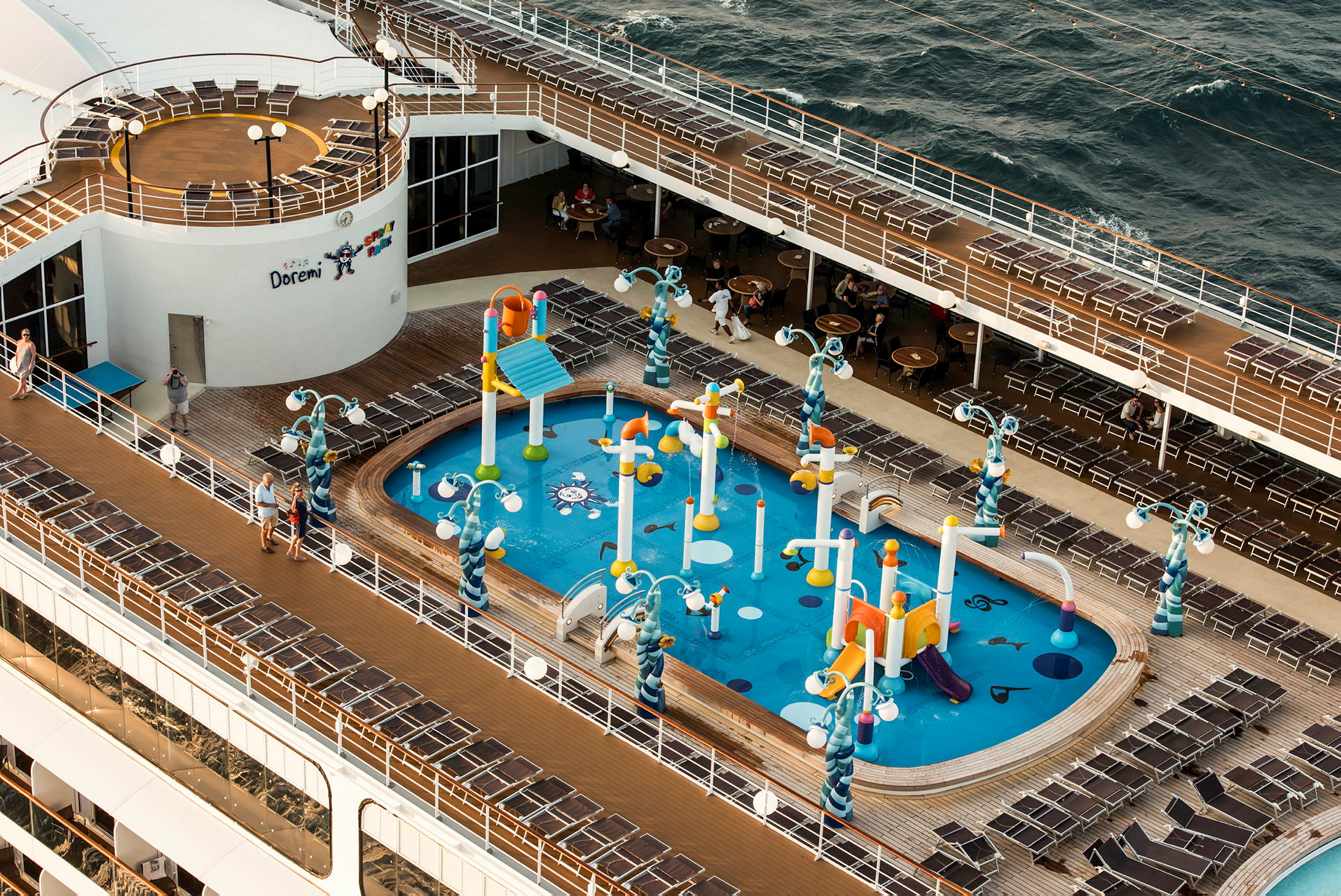 Bilde viser det øvre dekk på et cruiseskip med en vannlekeplass for barn og omkringliggende solsenger. (AI-generert tekst)