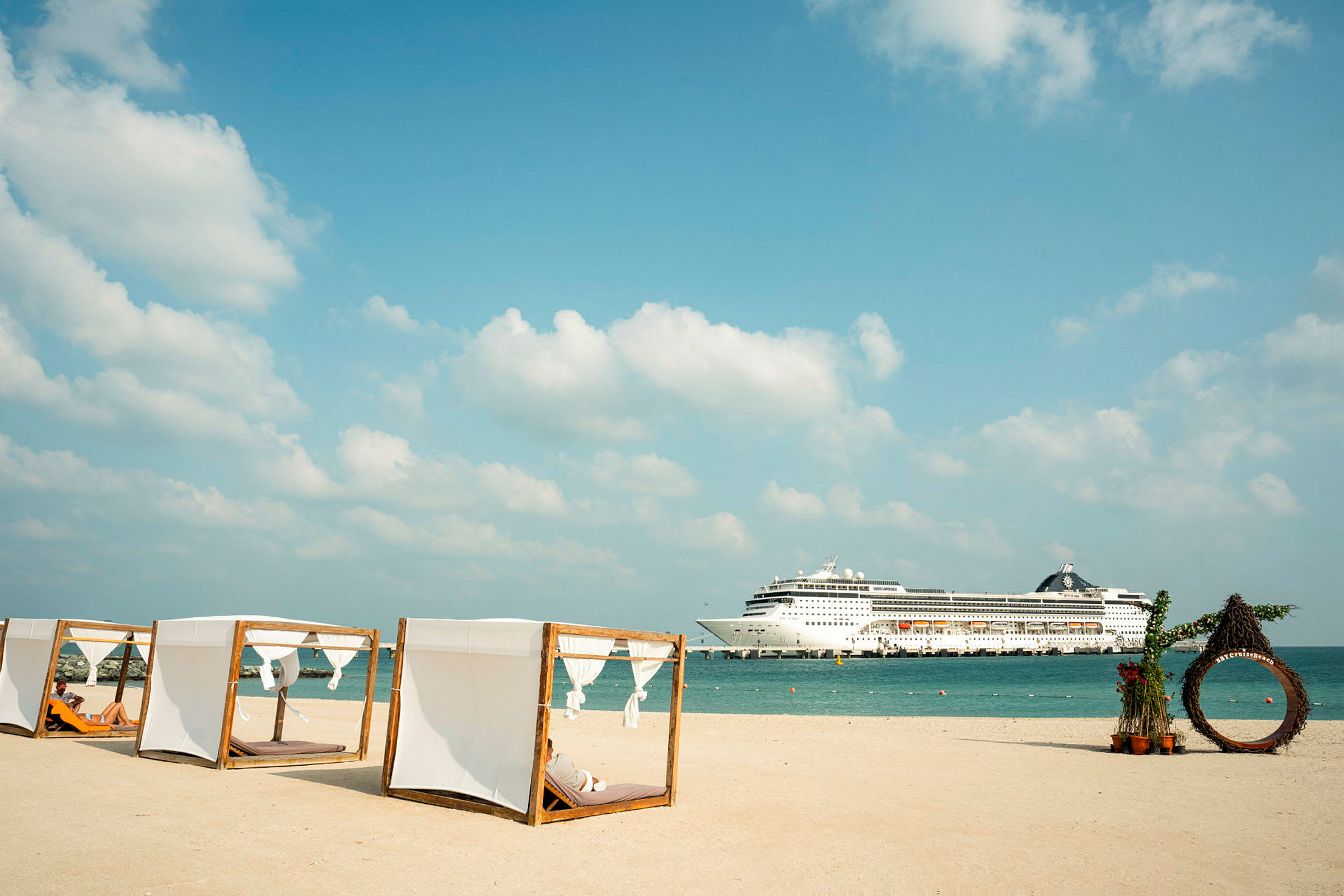 Bilde av en strand med skyggepaviljonger og et cruiseskip i bakgrunnen. (AI-generert tekst)