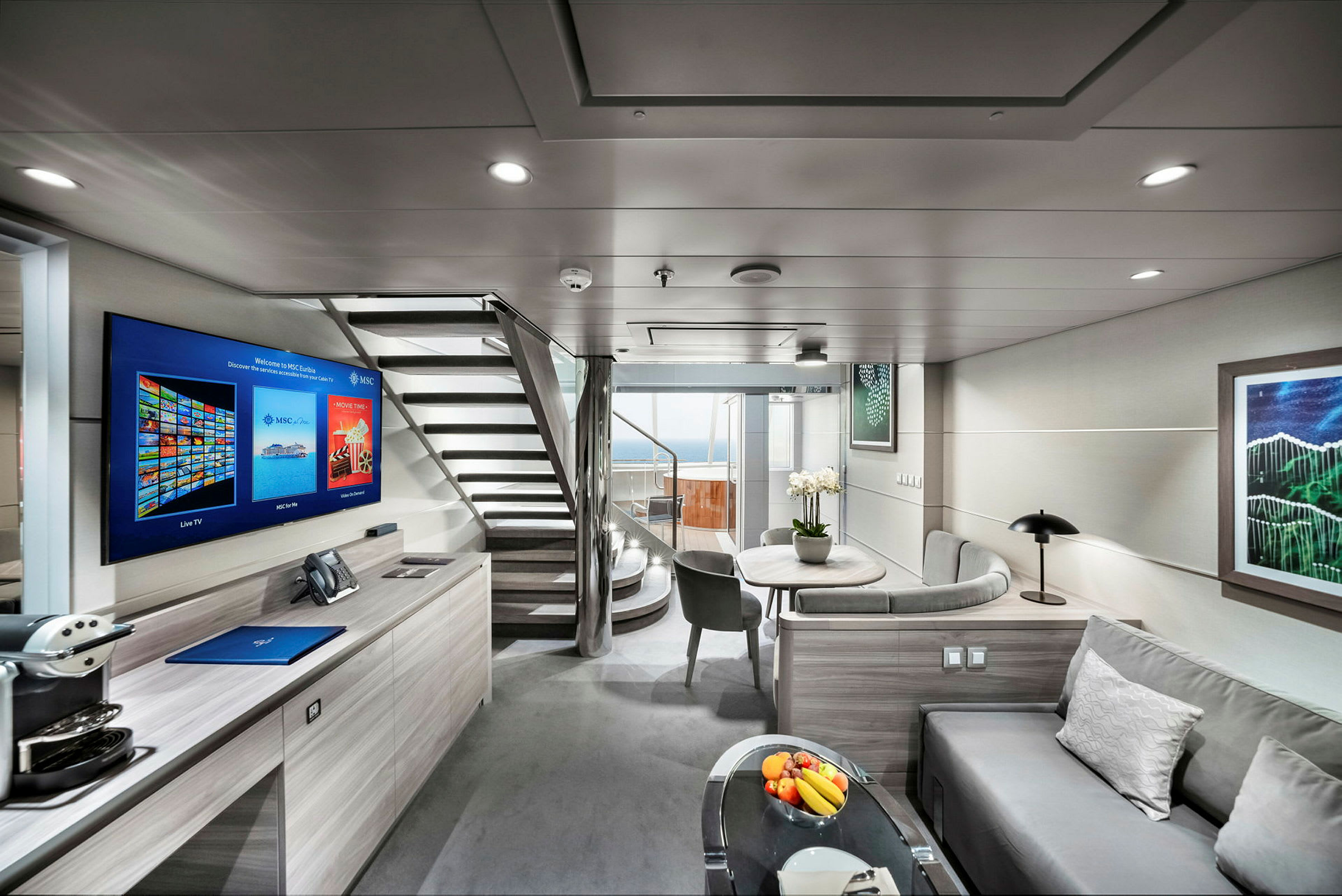 Et moderne og lyst interiør i en hotellsuite med sofa, bord, flatskjerm-TV og trapp som fører til en øvre etasje samt en balkong med utsikt. (AI-generert tekst)