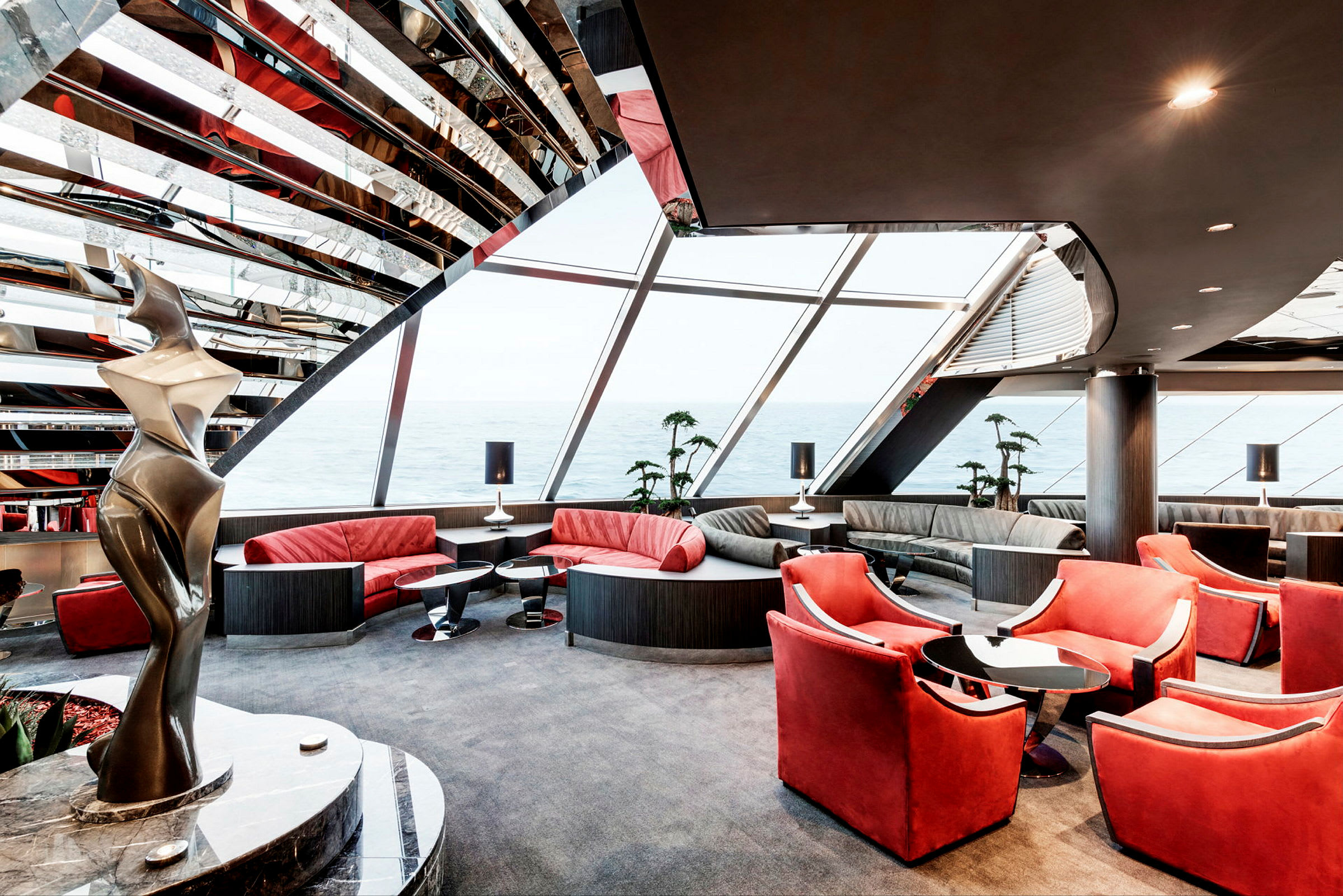 Et moderne innredet rom med røde stoler og sofaer, store vinduer med utsikt over havet og en abstrakt skulptur på siden. (AI-generert tekst)
