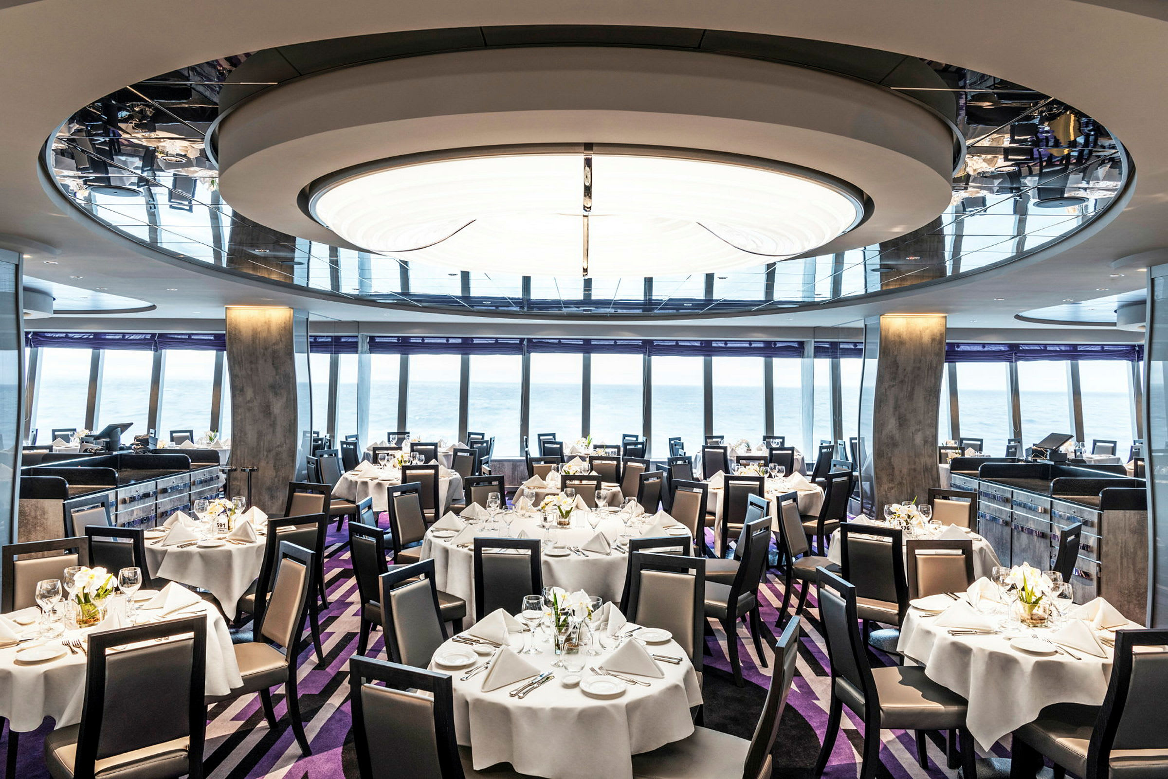Bilde av en elegant restaurant med bord dekket med hvite duker og stoler, vinduer med havutsikt ses i bakgrunnen. (AI-generert tekst)