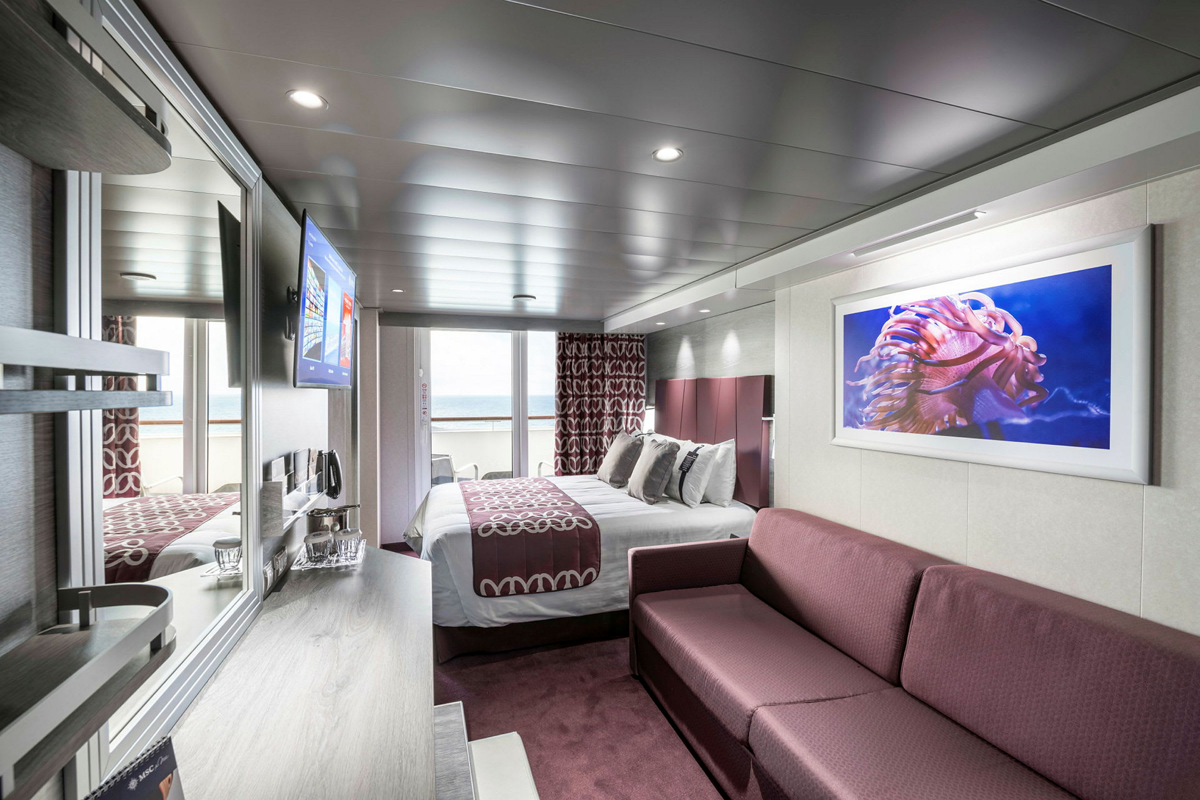Interiørbilde av et hotellrom med en seng, en sofa og havutsikt fra en balkong. (AI-generert tekst)