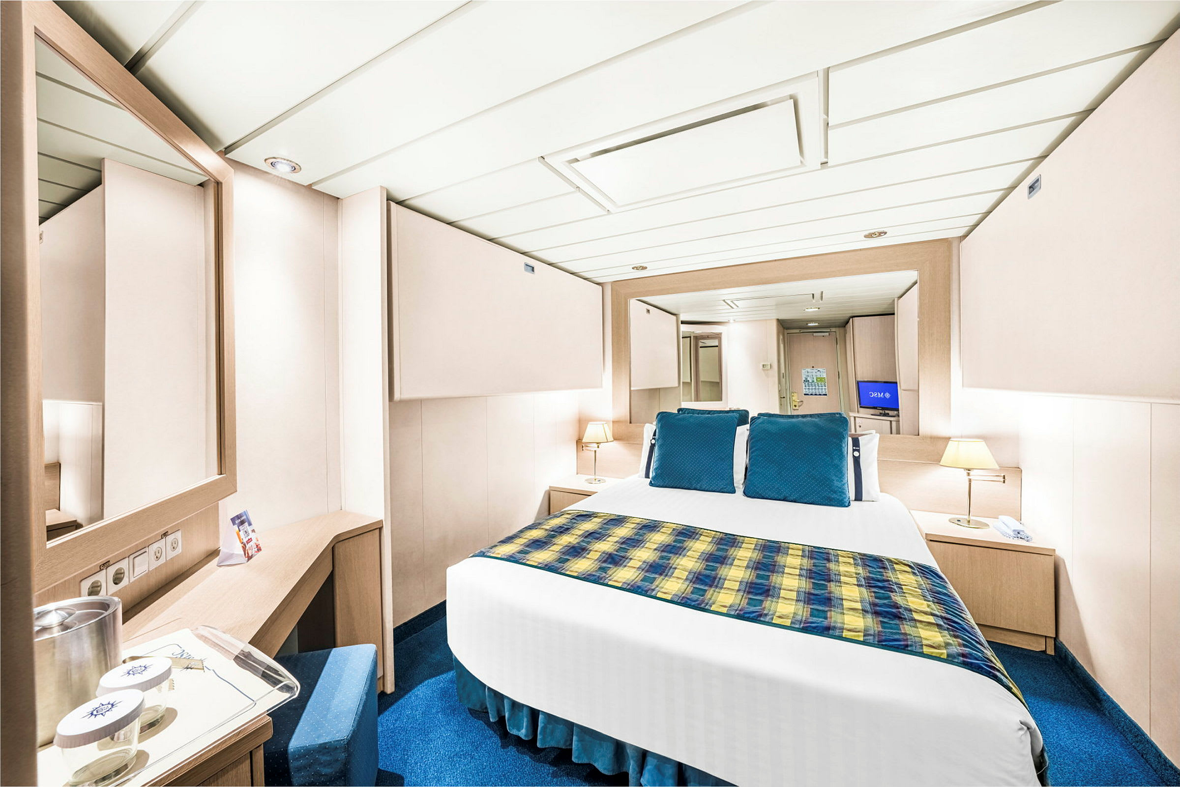 Et bilde av et hotellrom med moderne innredning, en seng med blå puter og gyllen mønstrett sengeteppe, to nattbord og speil. (AI-generert tekst)