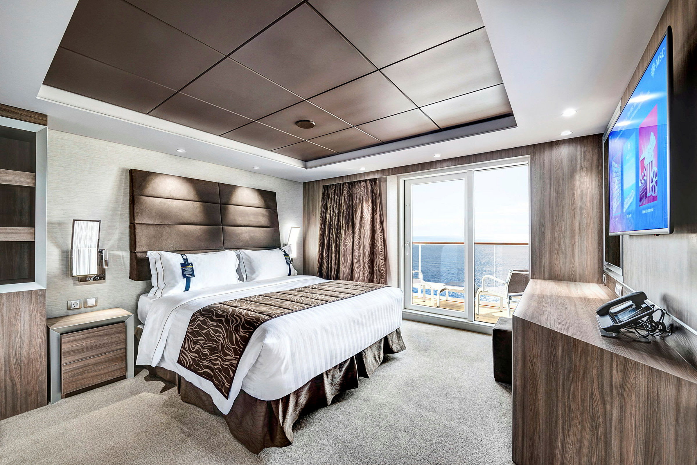 Et moderne hotellrom med en stor seng, en flatskjerm-TV, et skrivebord og en balkong med utsikt over havet. (AI-generert tekst)