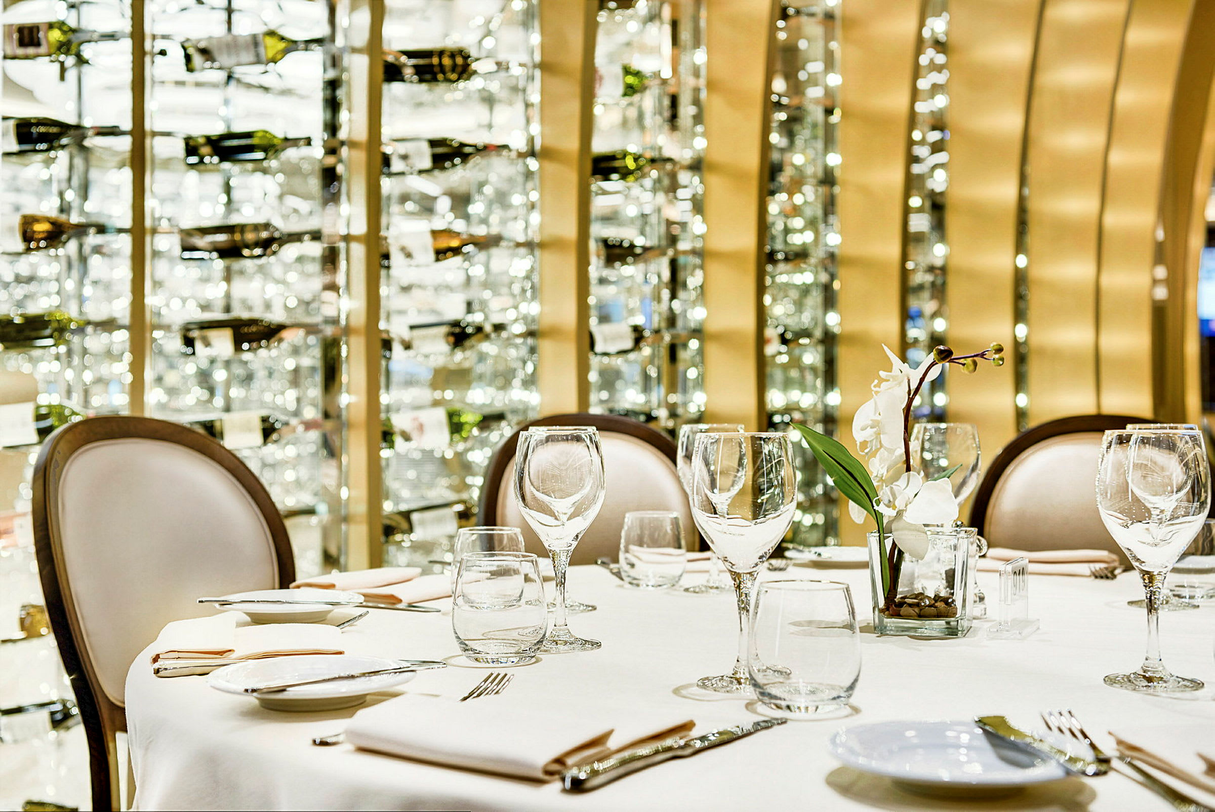 Bordsetting med glass, tallerkener og en hvit blomst på en restaurant med vinflasker i bakgrunnen. (AI-generert tekst)