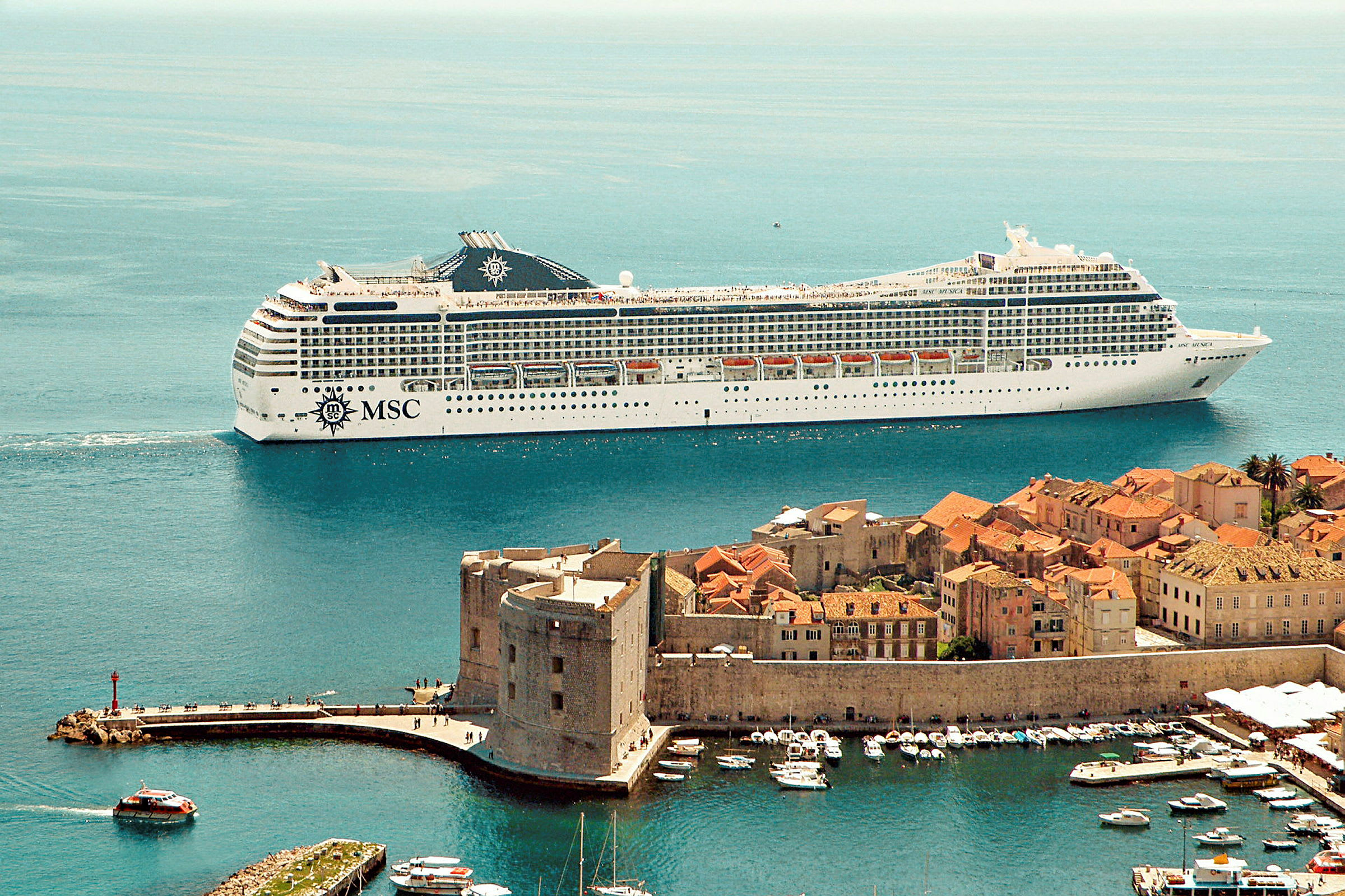 Bilde av et stort cruiseskip nær en historisk kystby med steinbygninger og havn. (AI-generert tekst)