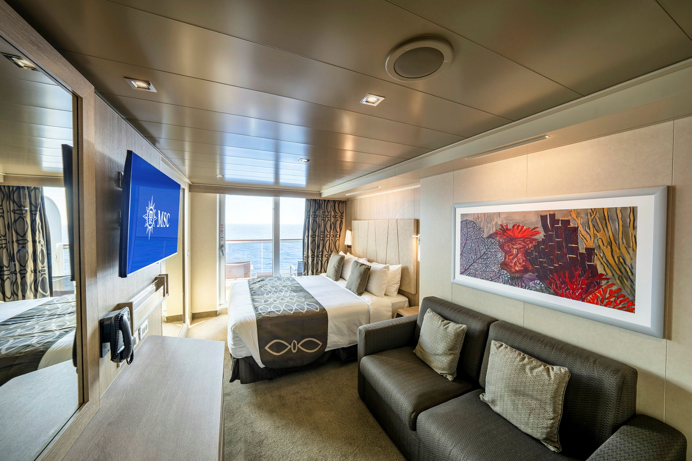 En moderne hotellsuite med havutsikt gjennom glassdører, inkludert en dobbeltseng, en sofa og en vegg-TV. (AI-generert tekst)