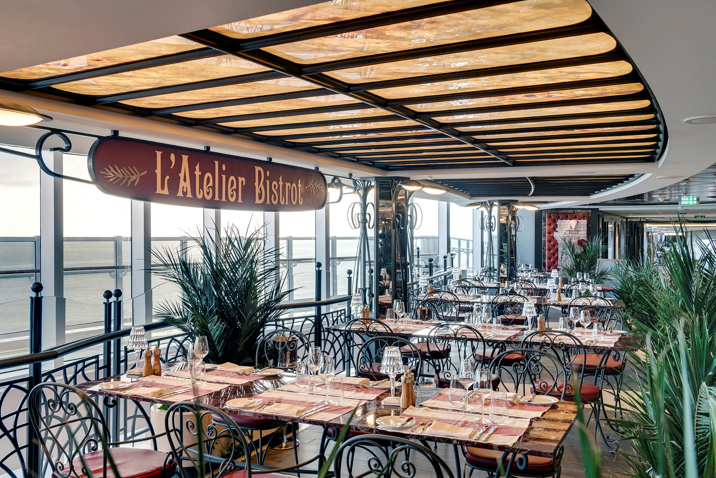 Utendørs serveringsområde til restauranten L'Atelier Bistro med bord og stoler og utsikt over havet. (AI-generert tekst)