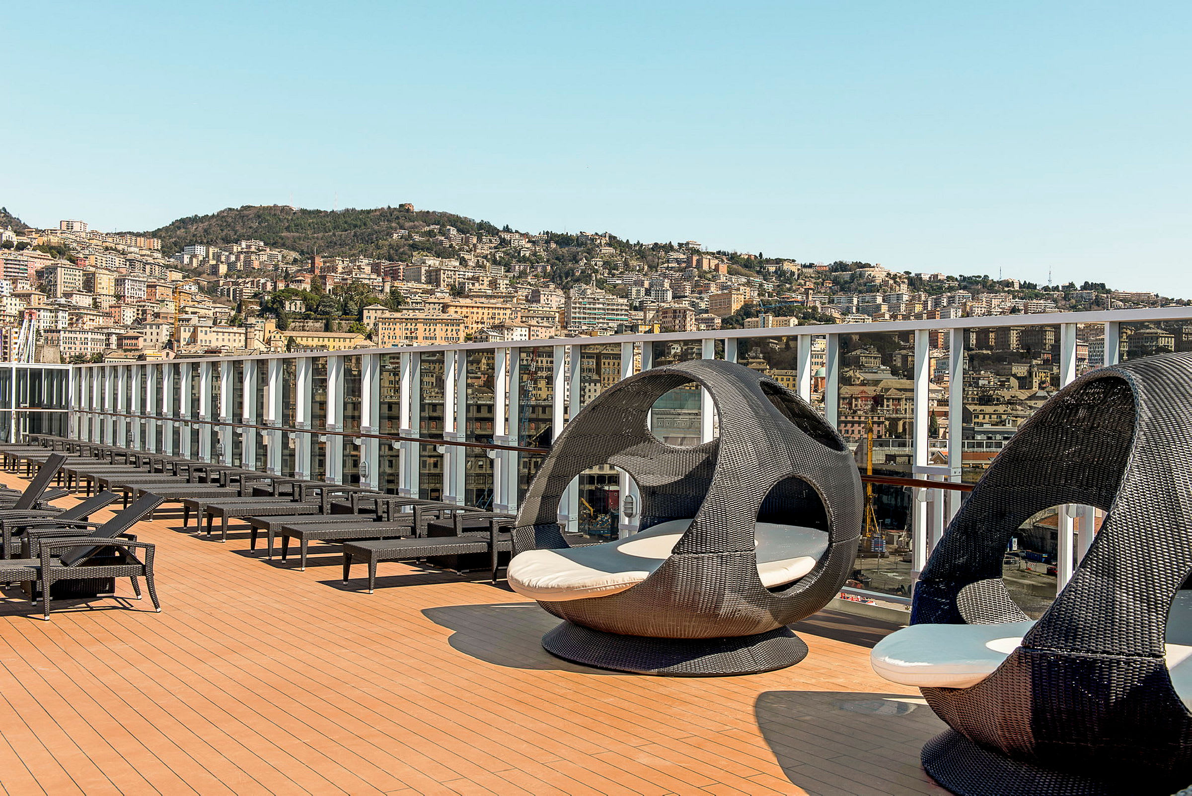 Bilde av en åpen terrasse med solsenger og runde rottingstoler. Bakgrunnen viser en by med åser og bygninger. (AI-generert tekst)